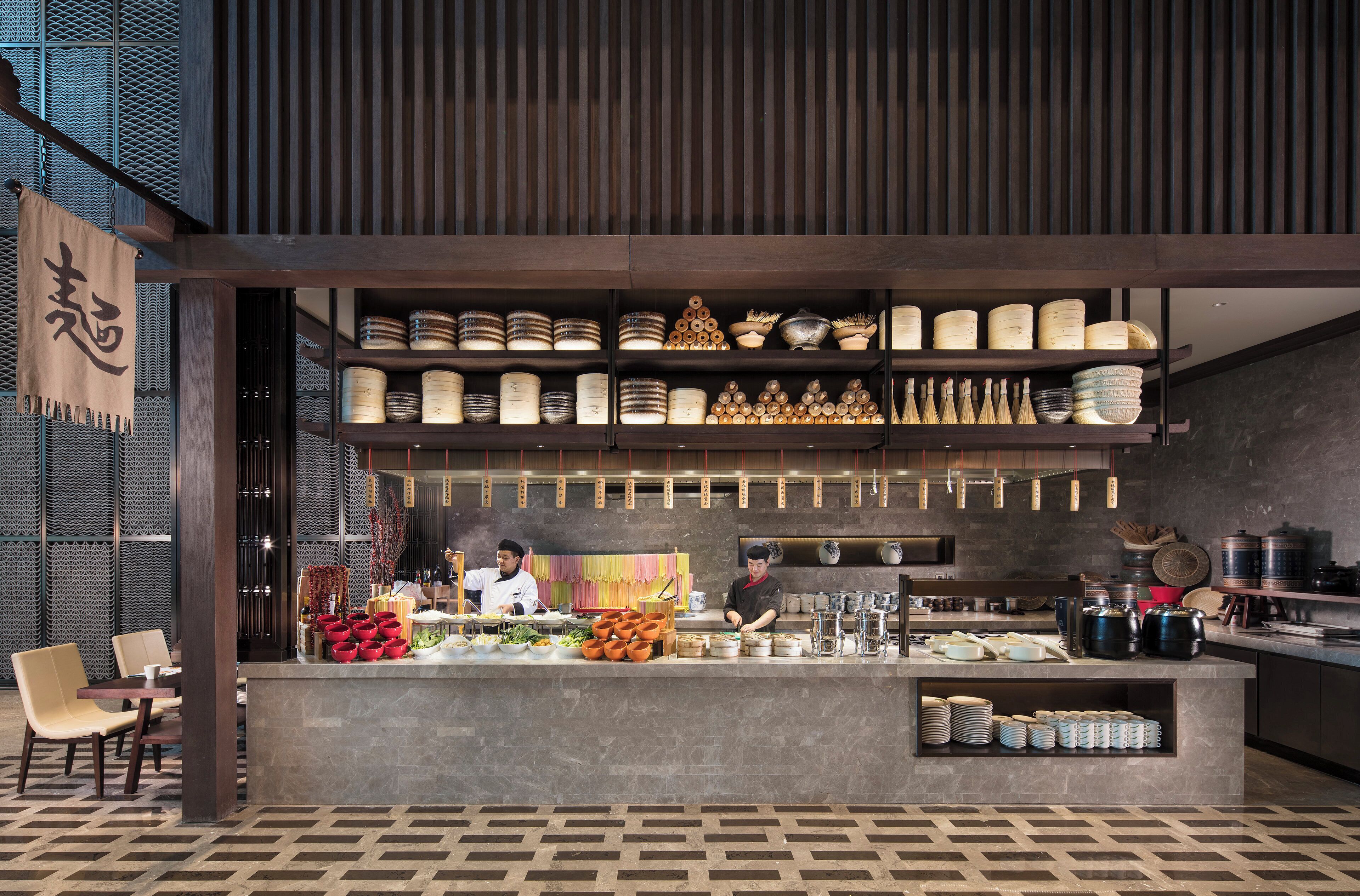 A restaurant interior with a central counter, shelves with dishes, and a chef preparing food.