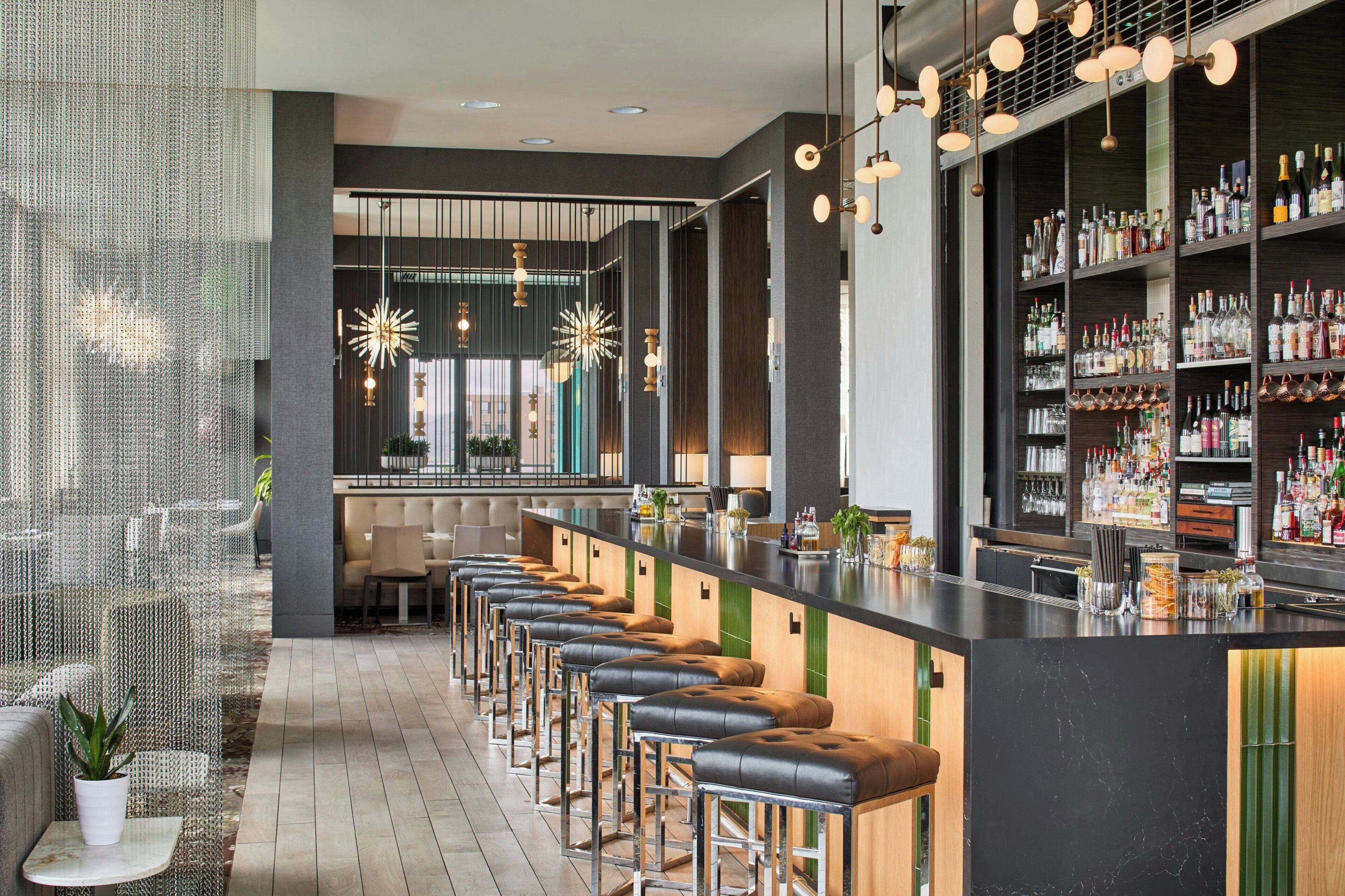 Un área de bar moderna con una barra larga, taburetes de cuero y una variedad de botellas y vasos en estantes.