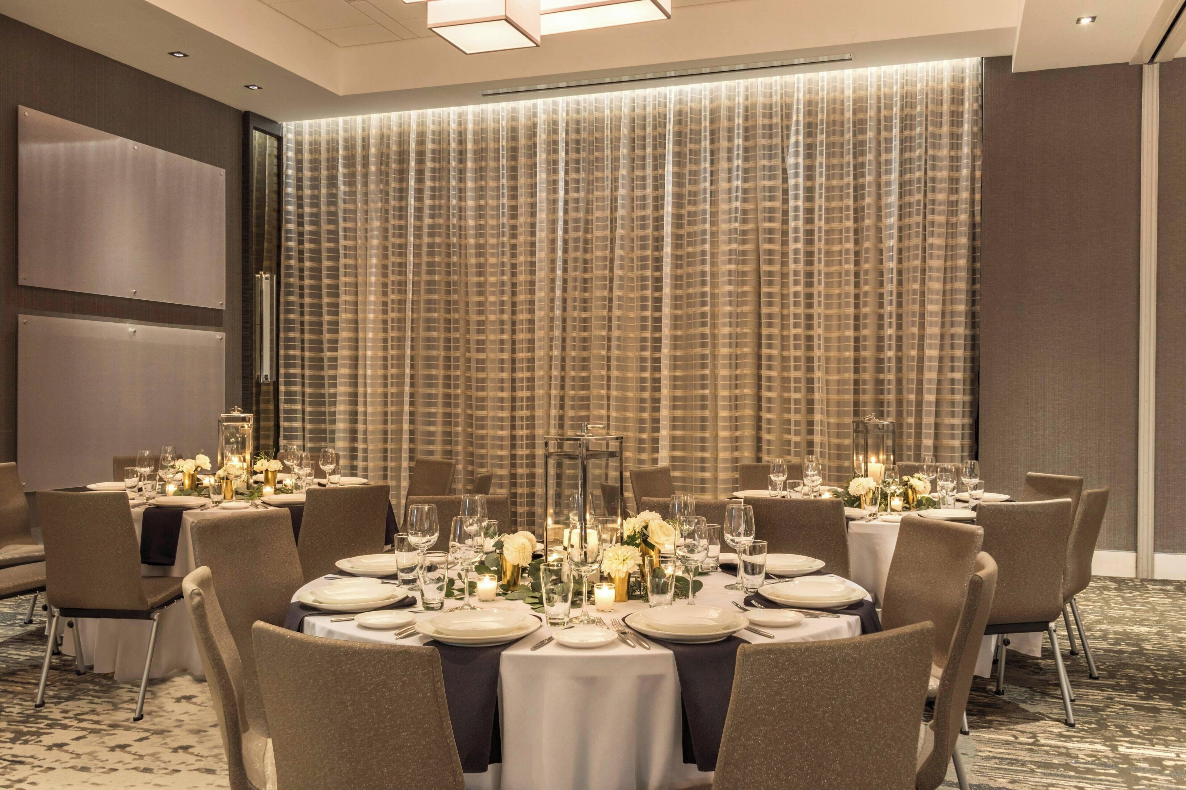 Un salón de banquetes con mesas redondas dispuestas para comer, destacando manteles blancos, velas y centros de mesa florales.