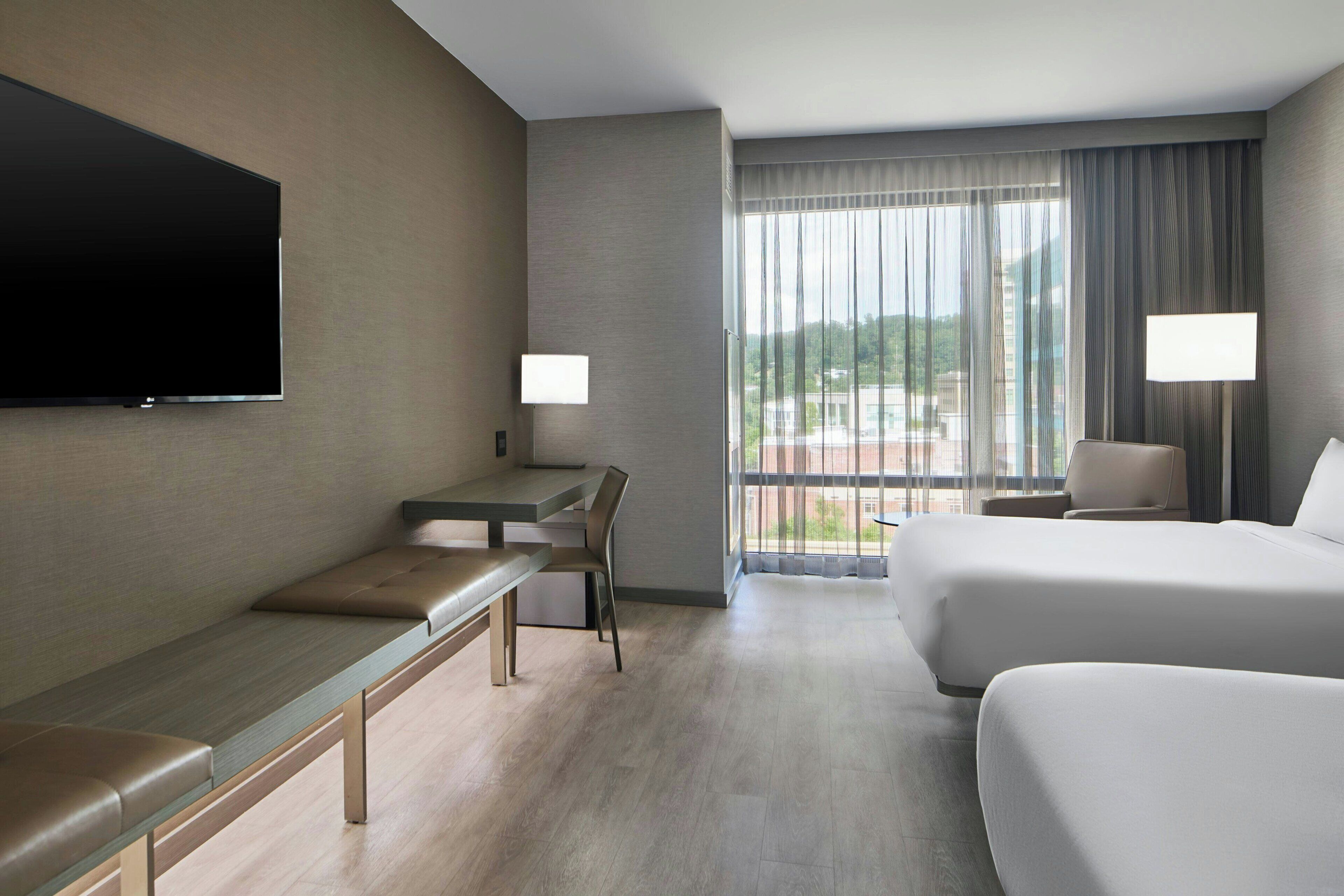 Habitación de hotel con televisión de pantalla plana, escritorio, silla, cama, sofá y un ventanal con cortinas.