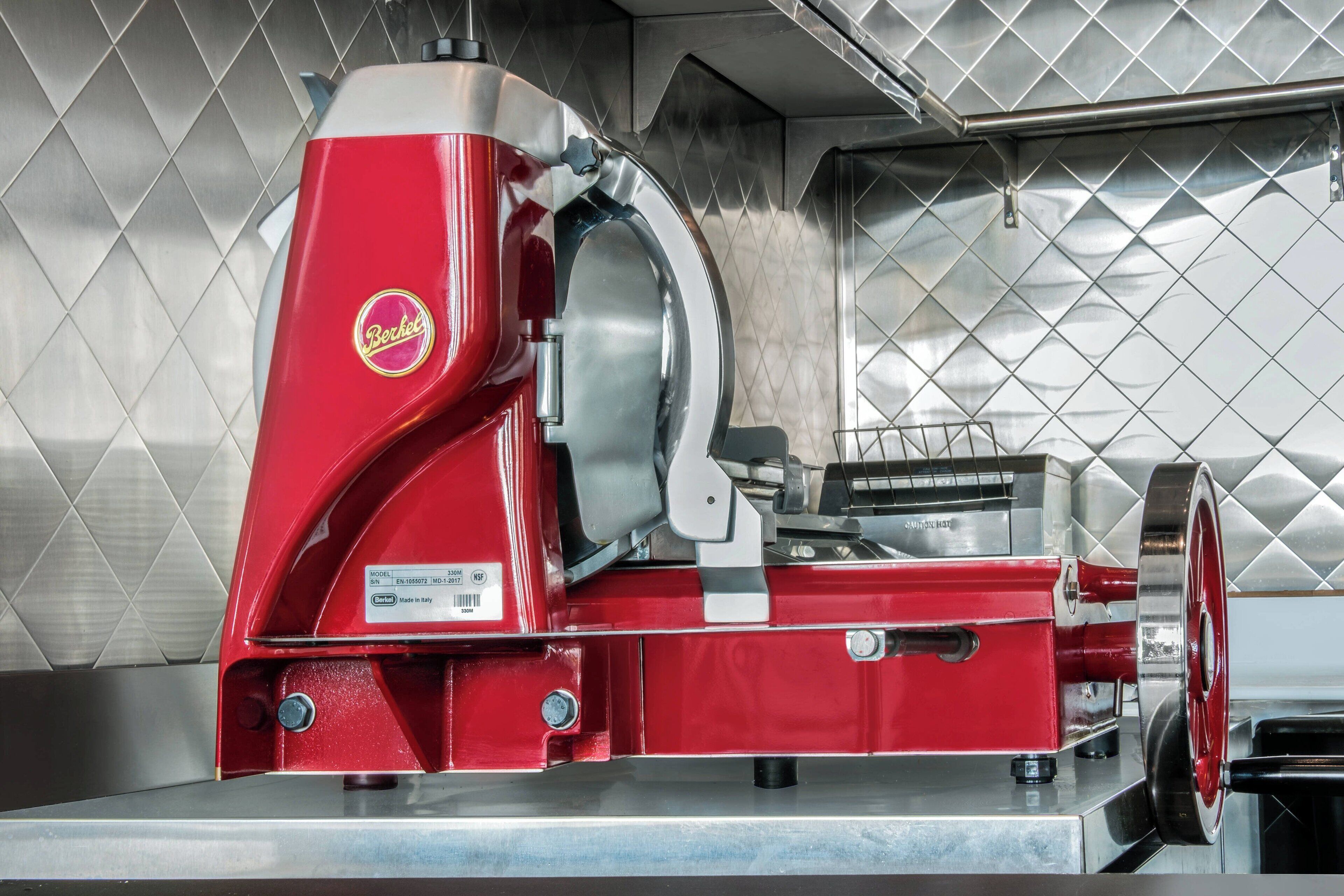 A red Berkel meat slicer in a kitchen with a diamond-patterned backsplash.