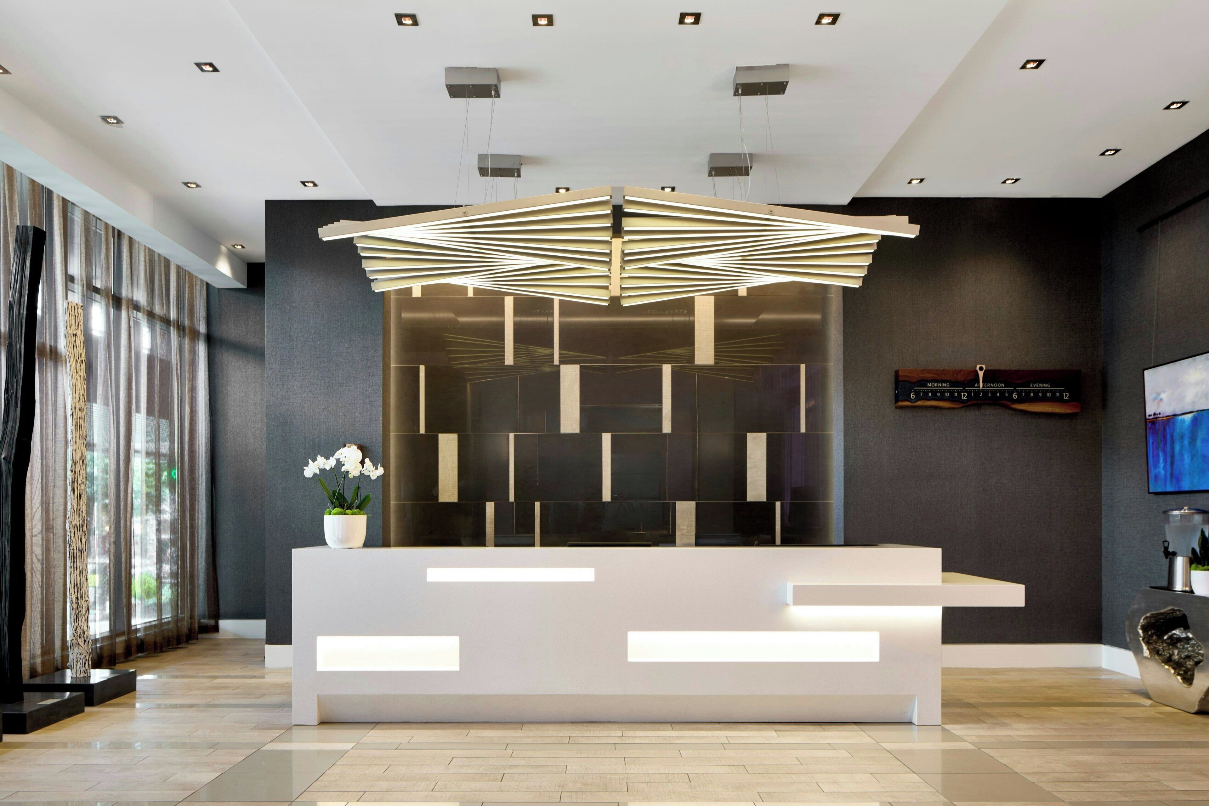 A modern reception desk with a geometric light fixture above it, a wall-mounted clock, and a potted orchid on the counter.