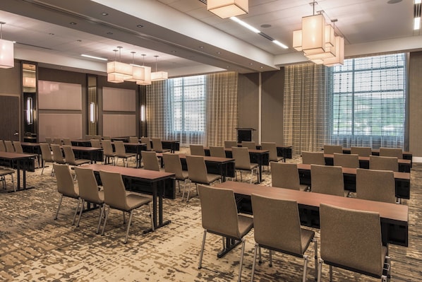 A conference room with rows of tables and chairs, large windows with blinds, and a carpeted floor.