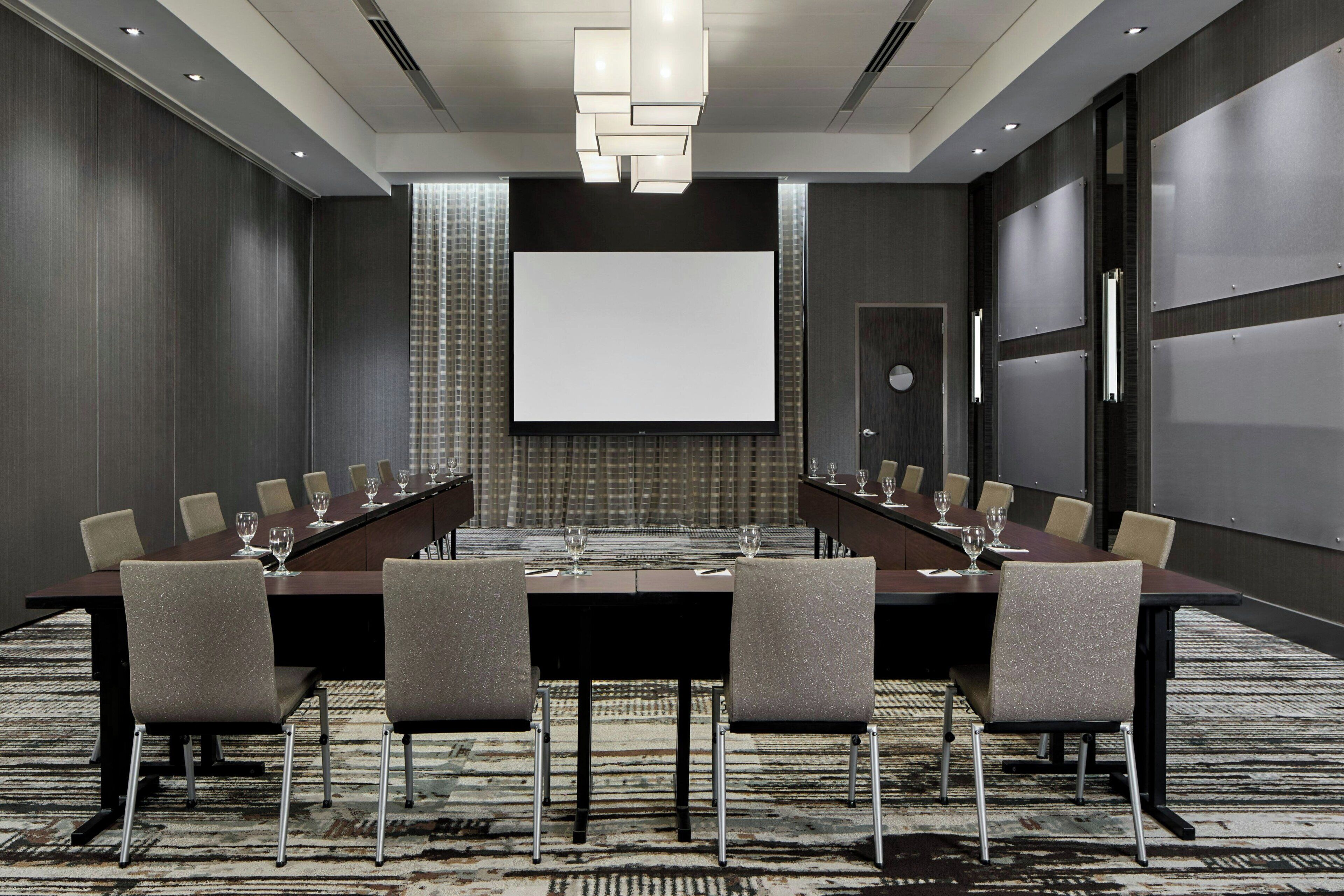 A conference room with a long table, chairs, a projector screen, and a carpeted floor.