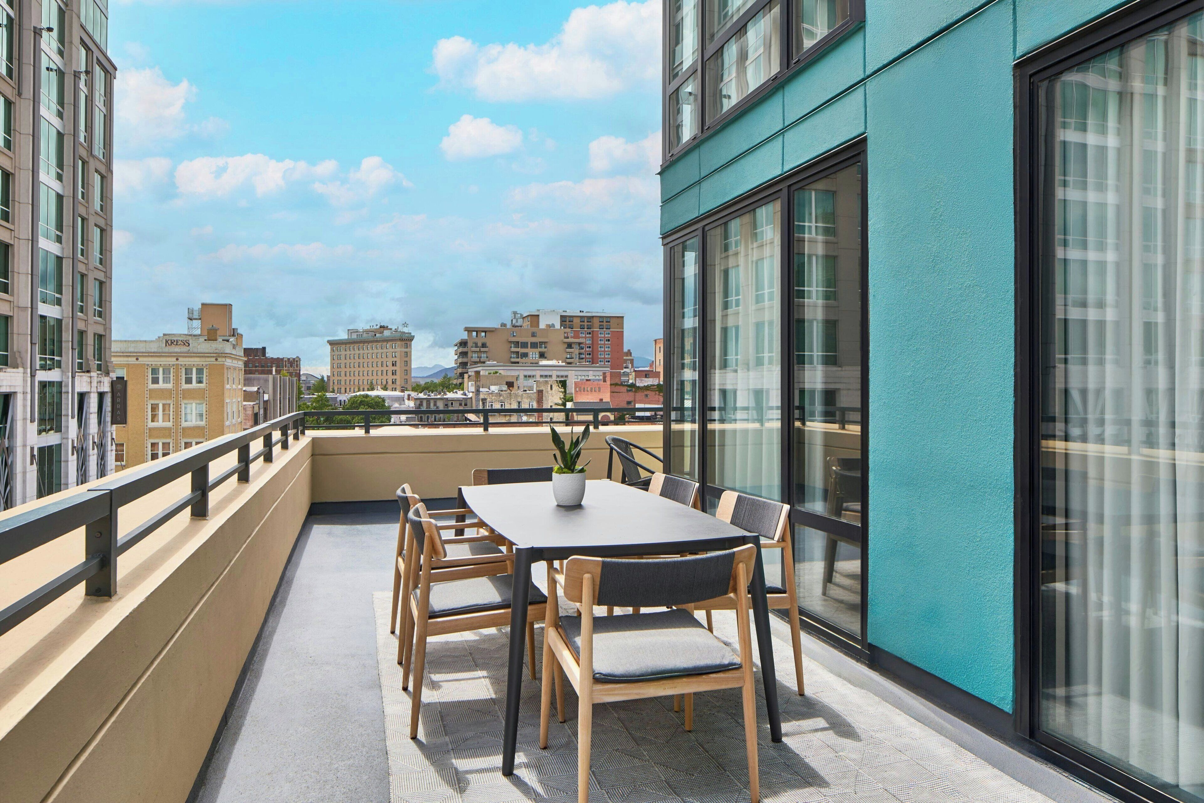 Un balcón con una mesa y sillas, una planta en maceta y vistas de edificios cercanos.