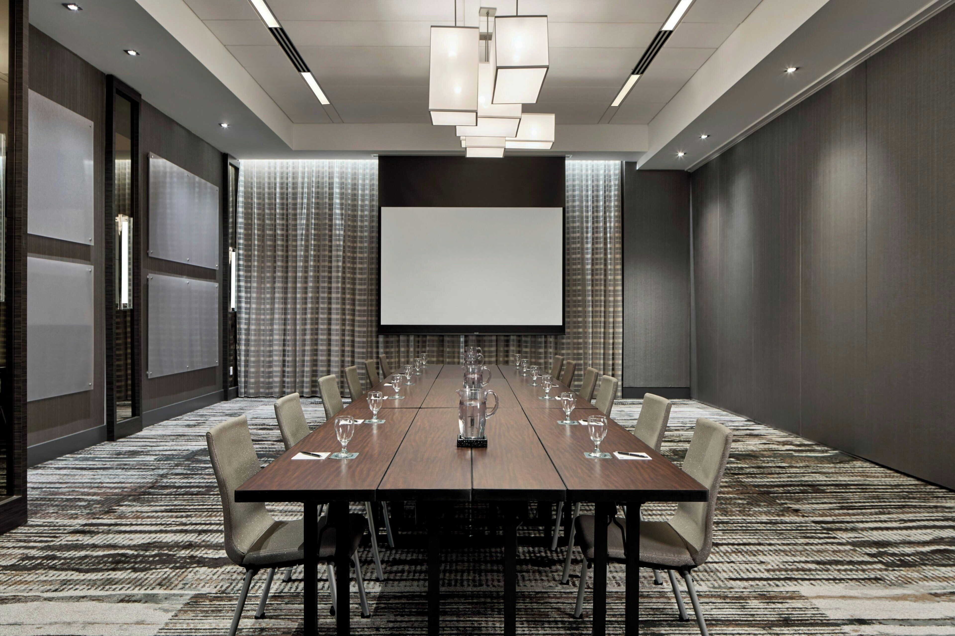 A conference room with a long table, chairs, a projector screen, and a patterned carpet.