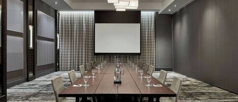 A conference room with a long table, chairs, a projector screen, and a patterned carpet.