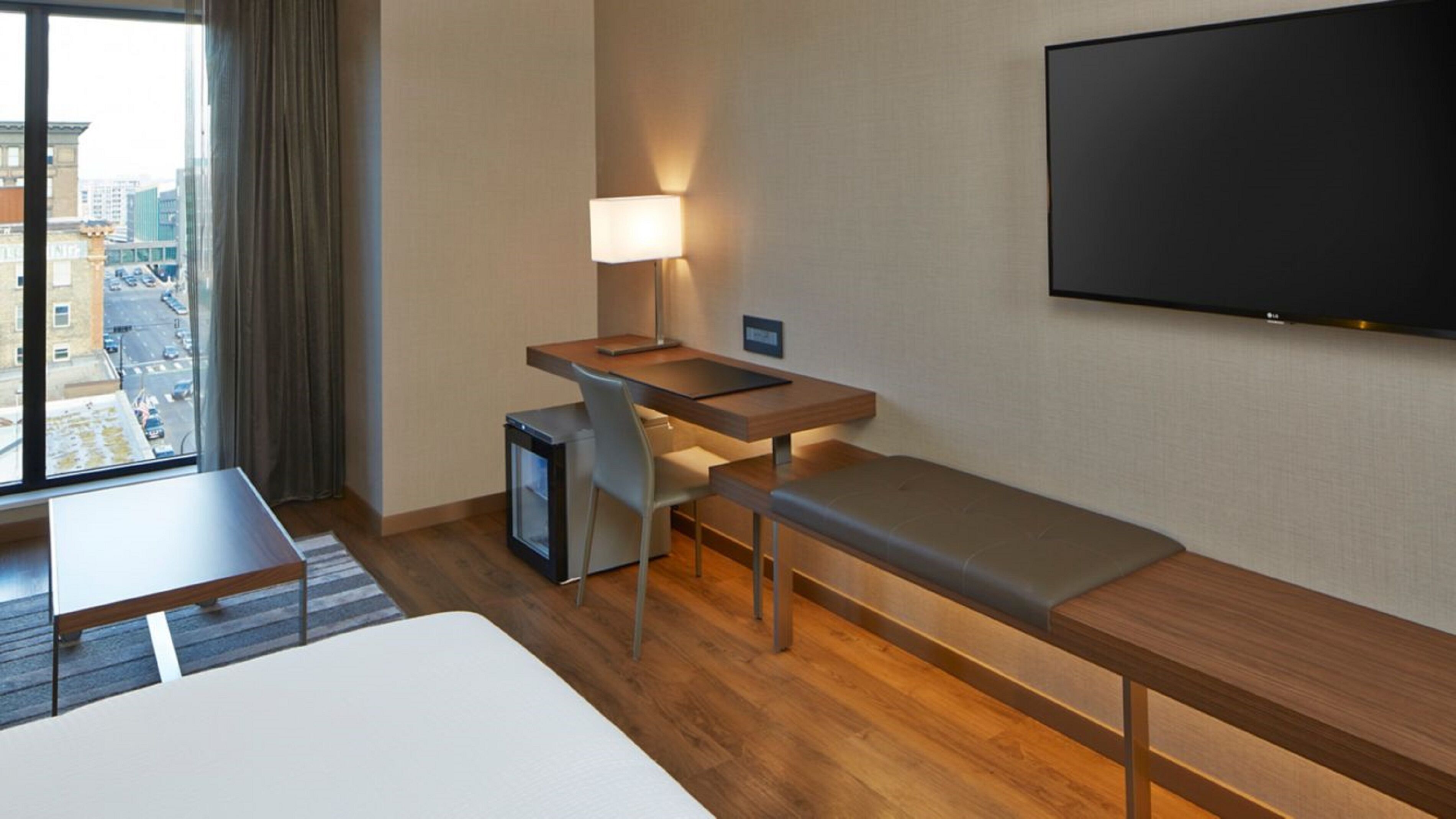 Une chambre d’hôtel comprenant un lit, un bureau avec une chaise, une petite télévision, un banc, une lampe et un téléviseur fixé au mur.