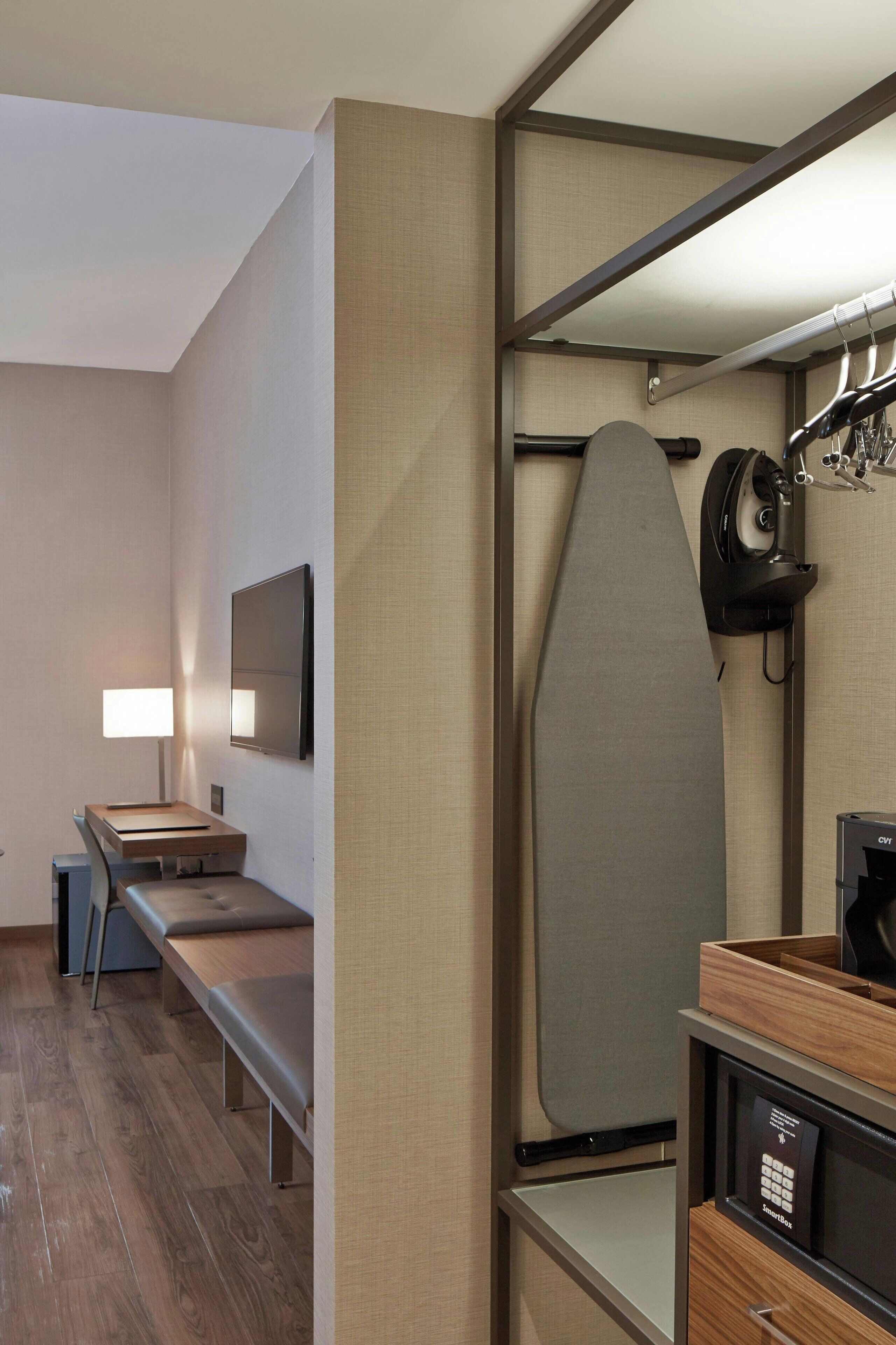 A modern hotel room with a flat-screen TV, a bench, a wooden side table, a built-in ironing board, and a coffee machine.