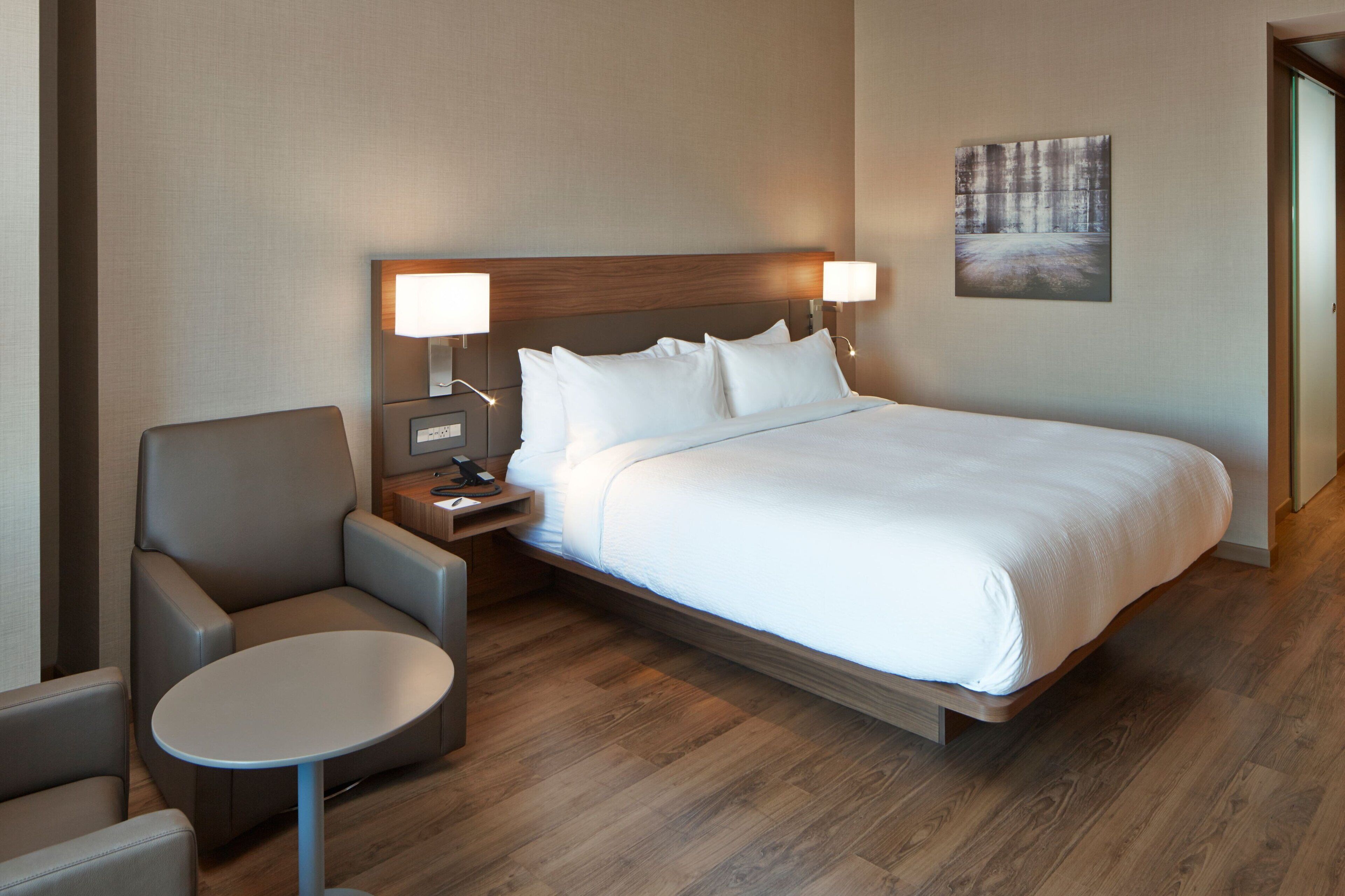 Hotelkamer met een bed, een stoel, een klein tafeltje, een nachtkastje met een lamp en een schilderij aan de muur.