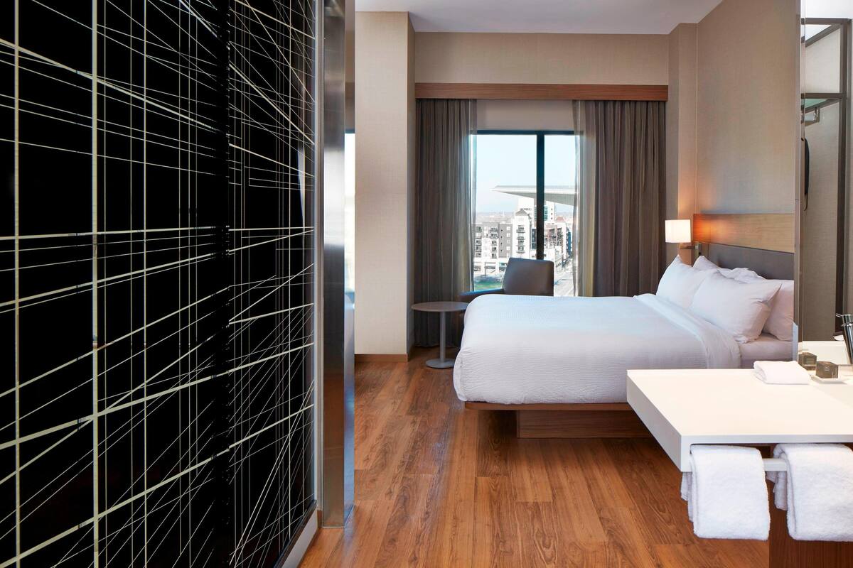 Habitación de hotel con una cama grande, una mesita de noche con lámpara, una pequeña mesa redonda y vistas a la ciudad a través de amplias ventanas.
