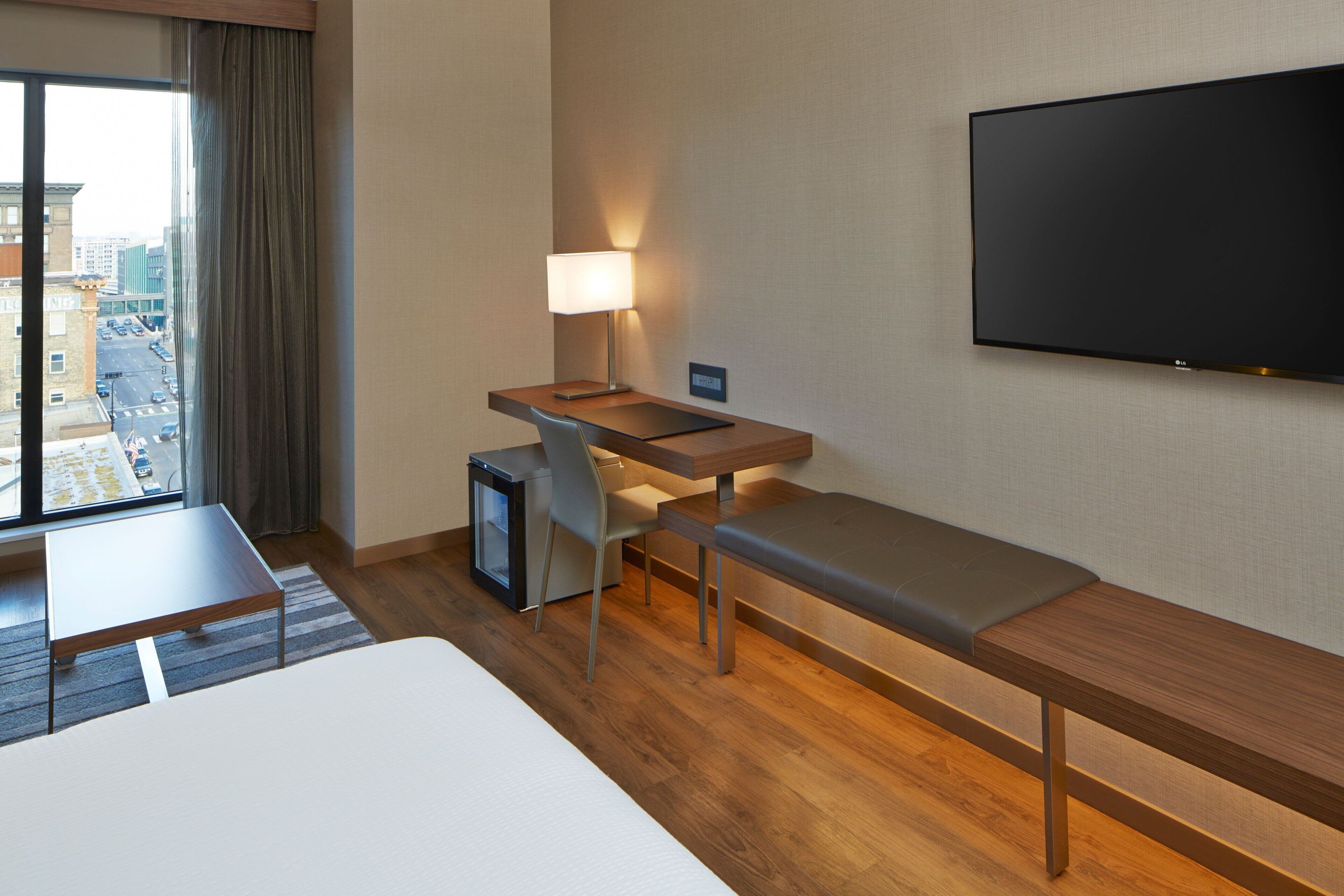 Ein Hotelzimmer mit großem Fenster, einem Schreibtisch mit Lampe, einem Fernseher, einem Bett und einer Bank.