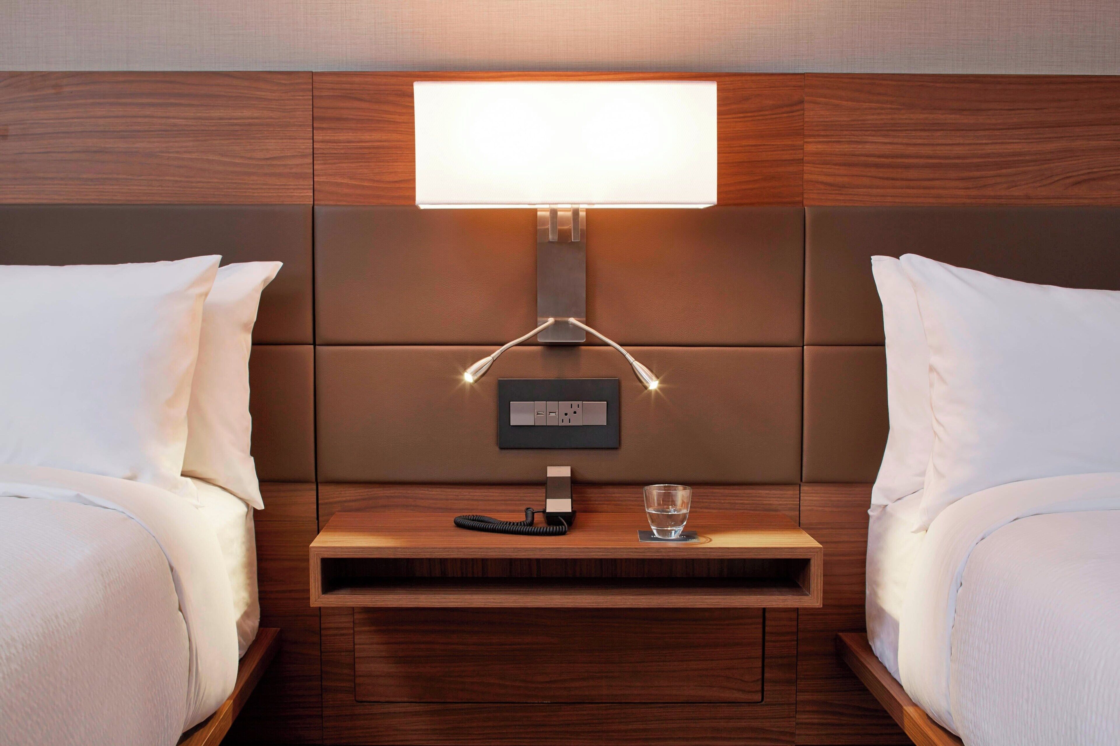 A hotel room with a wooden headboard, a bedside table with a lamp, a phone, and a glass of water.