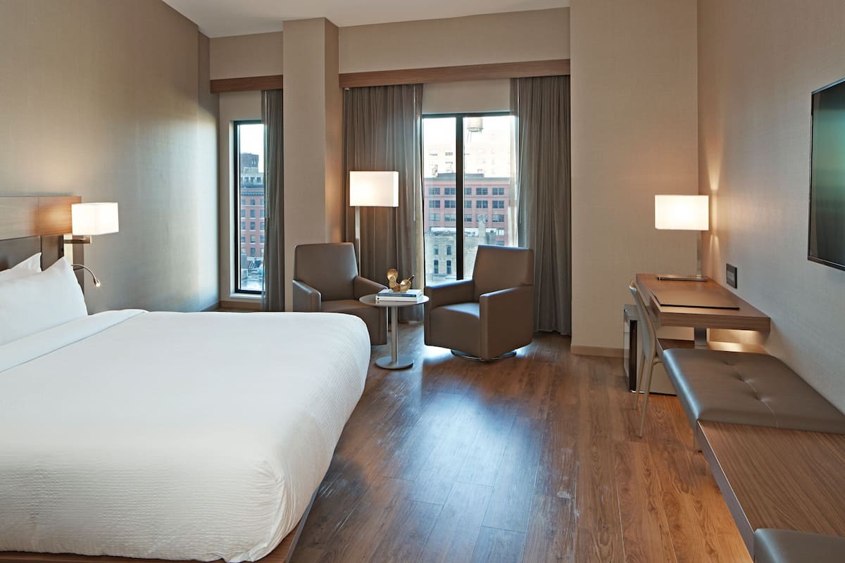 Habitación de hotel con una cama grande, dos sillones, una mesita y un escritorio con una silla. Hay dos ventanas con cortinas, y se aprecia un paisaje urbano a través de ellas.