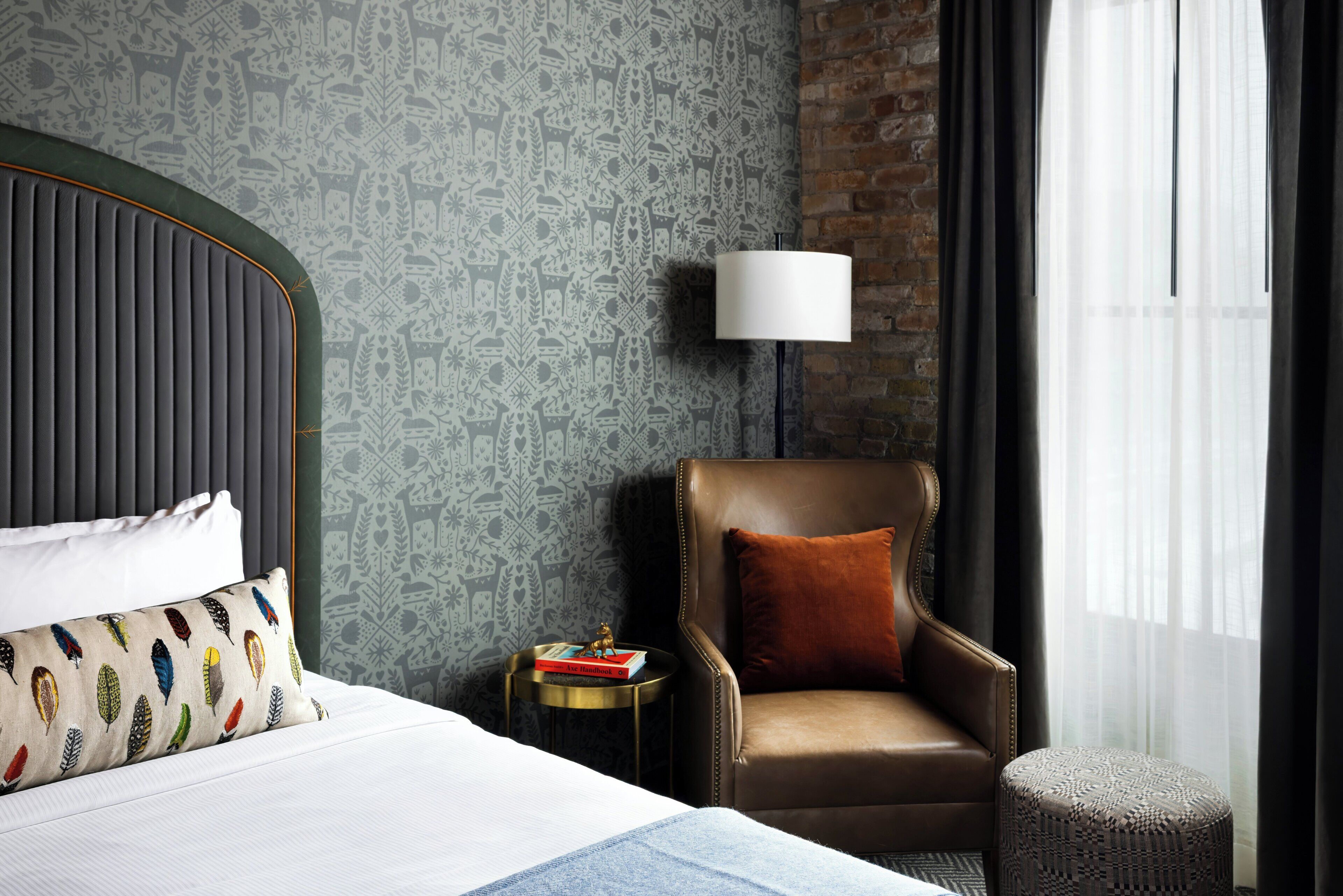 A hotel room with a bed, a leather armchair, a side table with books, a lamp, and a patterned wall.