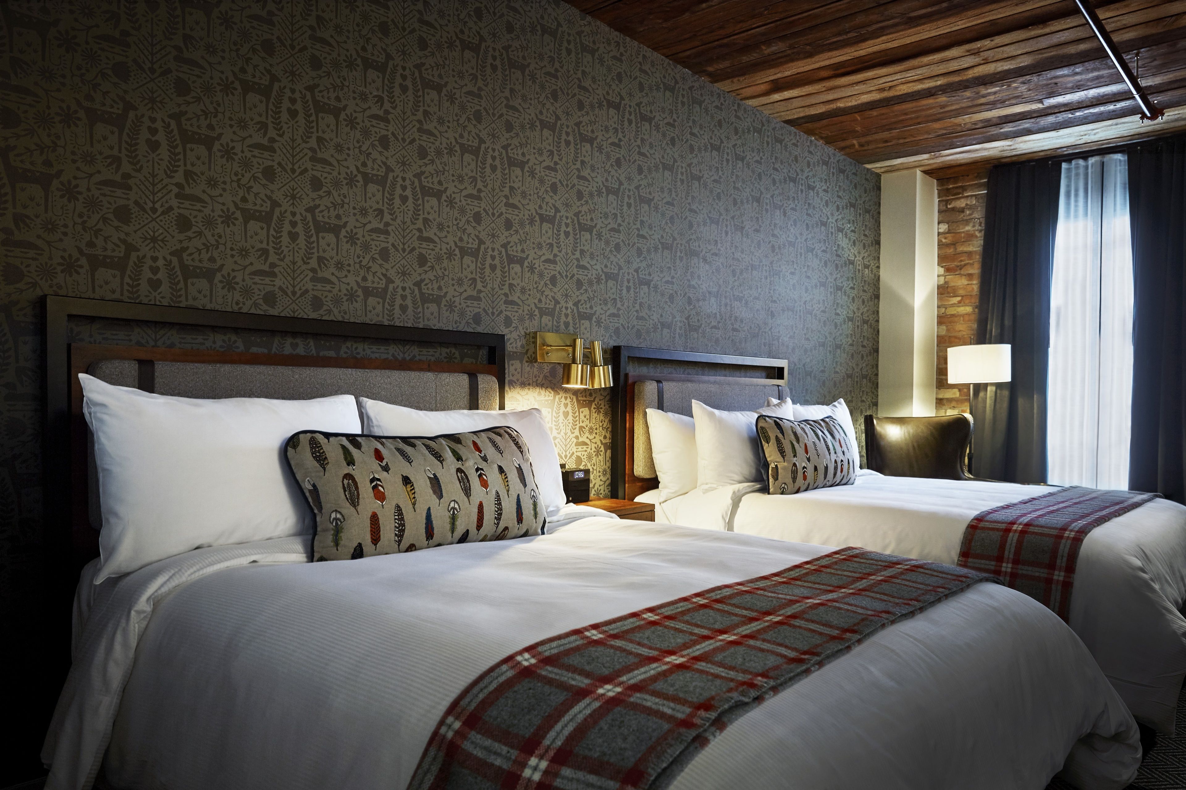 A hotel room with two beds, patterned wallpaper, a wooden ceiling, and a window with curtains.