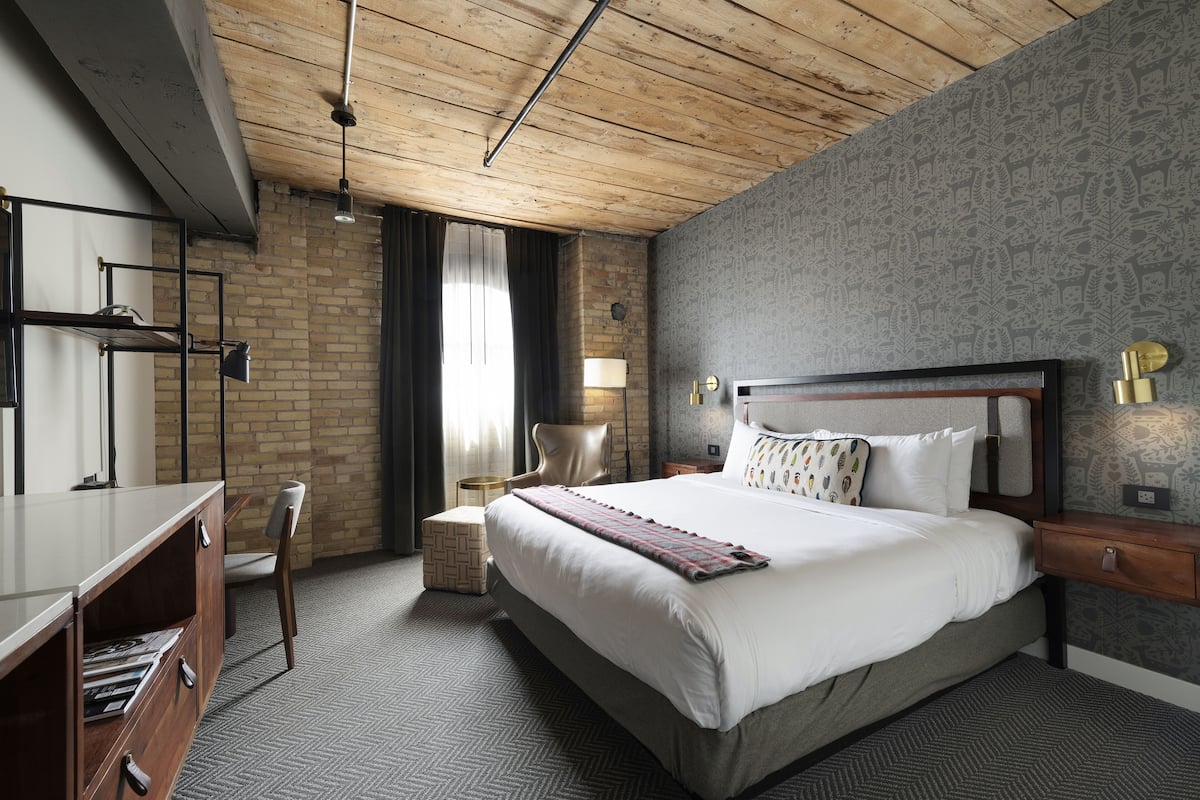 A modern hotel room with a large bed, a wooden ceiling, a brick wall, and a patterned wallpaper.