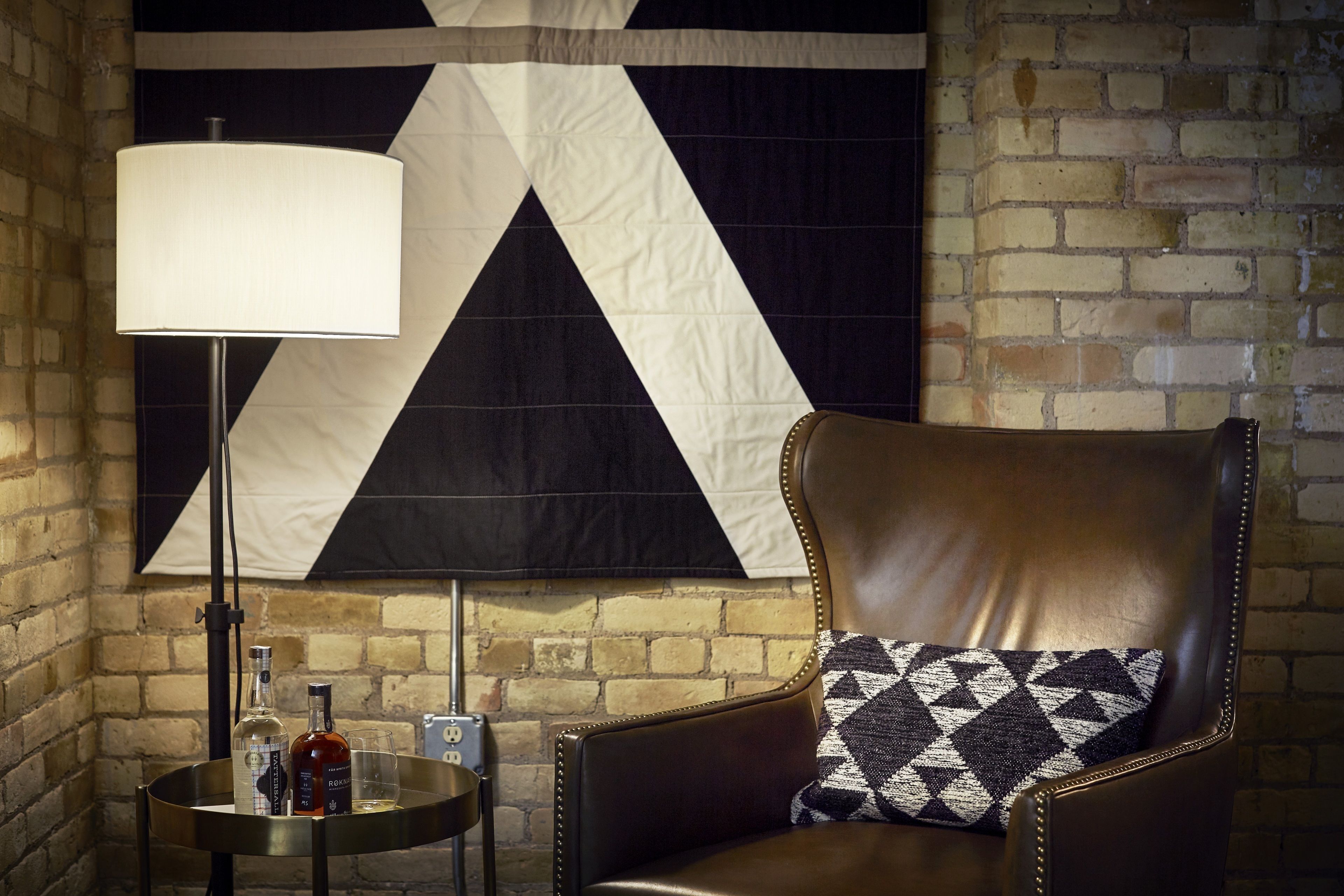 A leather armchair with a patterned pillow, a table with bottles and glasses, and a large wall tapestry.