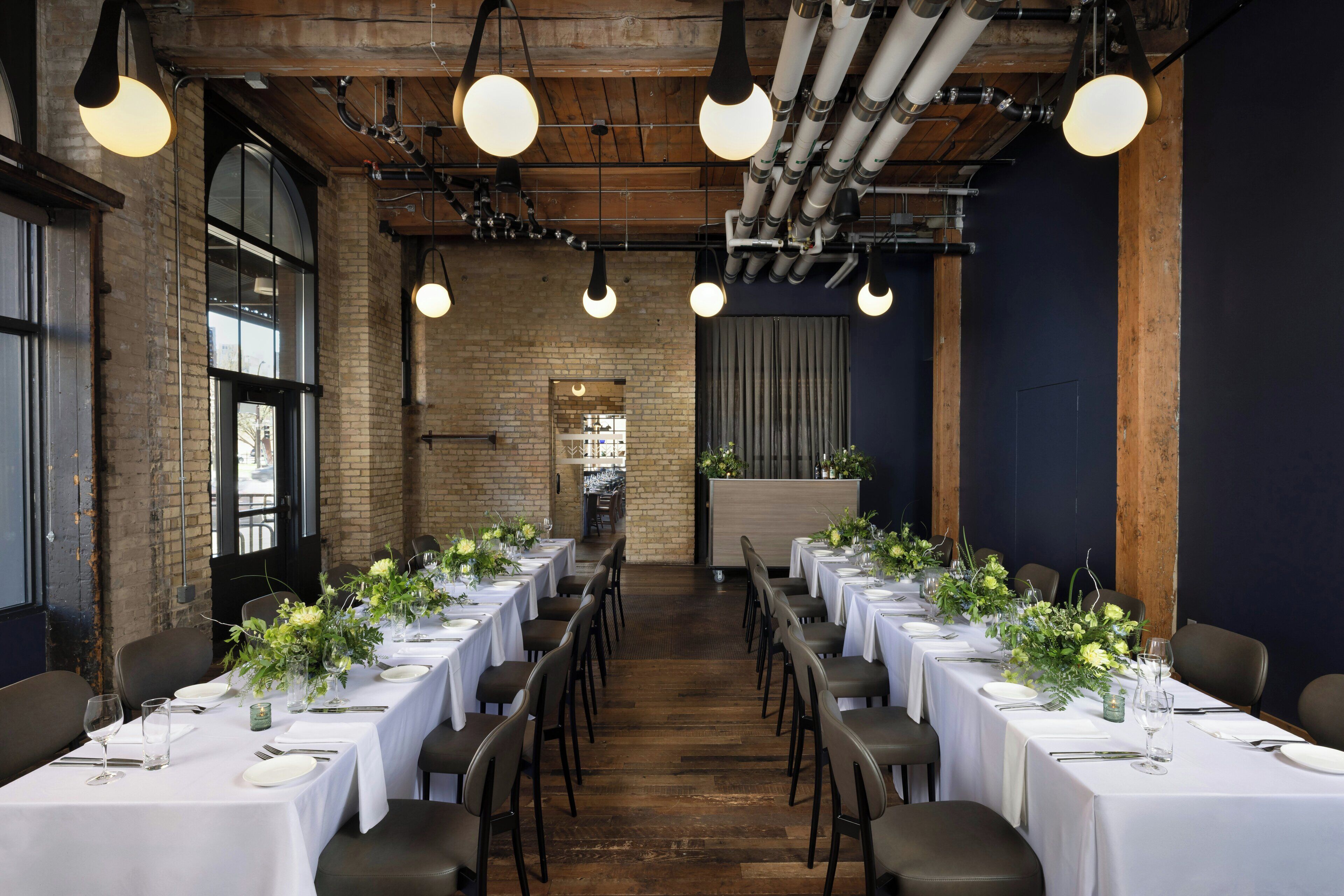 A long dining table with white tablecloths, chairs, and floral centerpieces, set in a room with exposed brick walls and wooden beams.