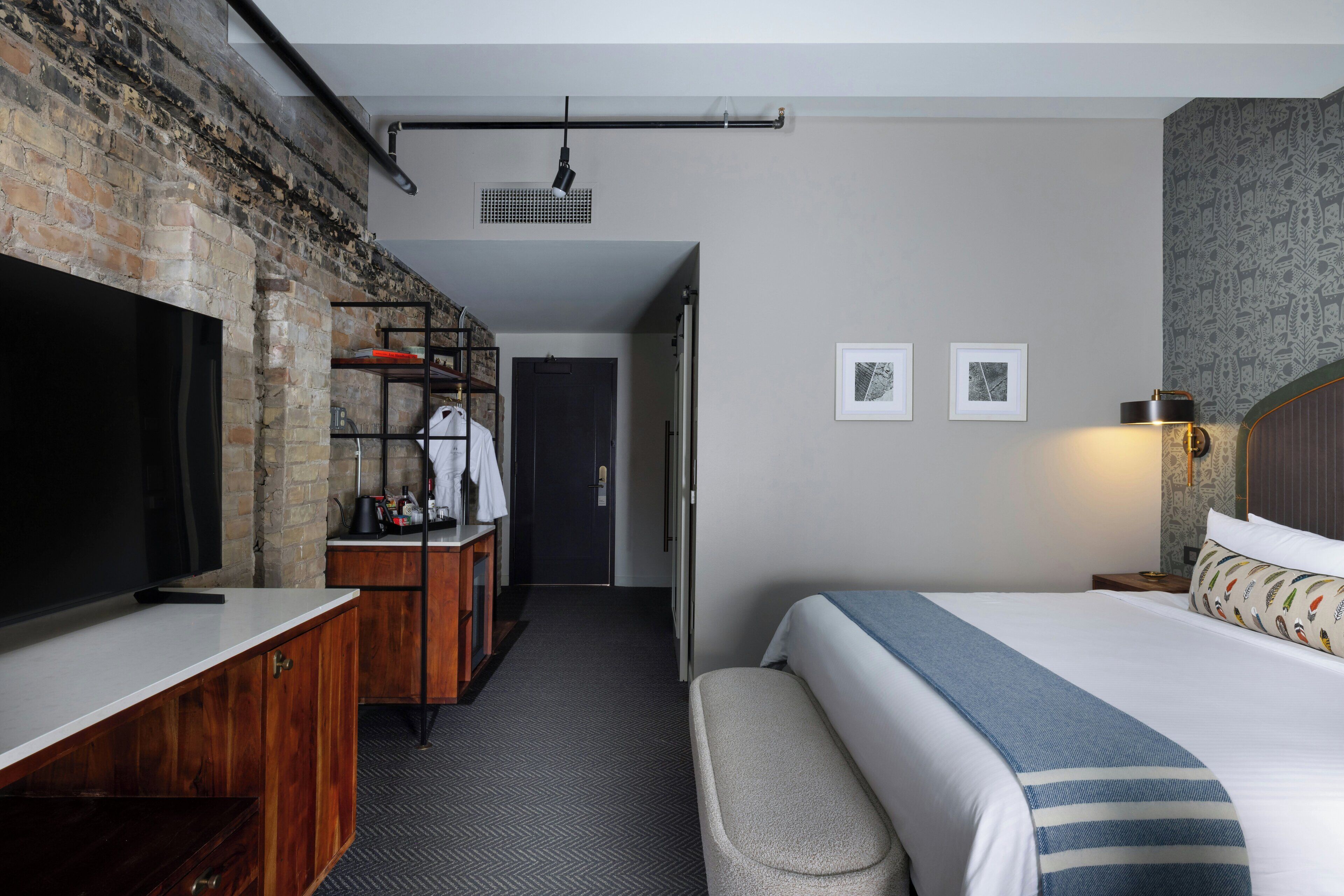 Habitación de hotel con pared de ladrillo, televisor de pantalla plana, cómoda de madera, cama con manta a rayas y mesita de noche con lámpara.