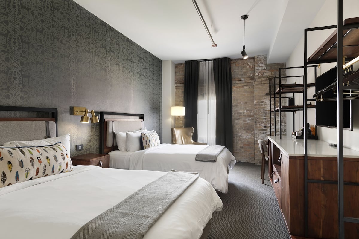 A hotel room with two beds, a desk, a TV, and a large window with curtains.