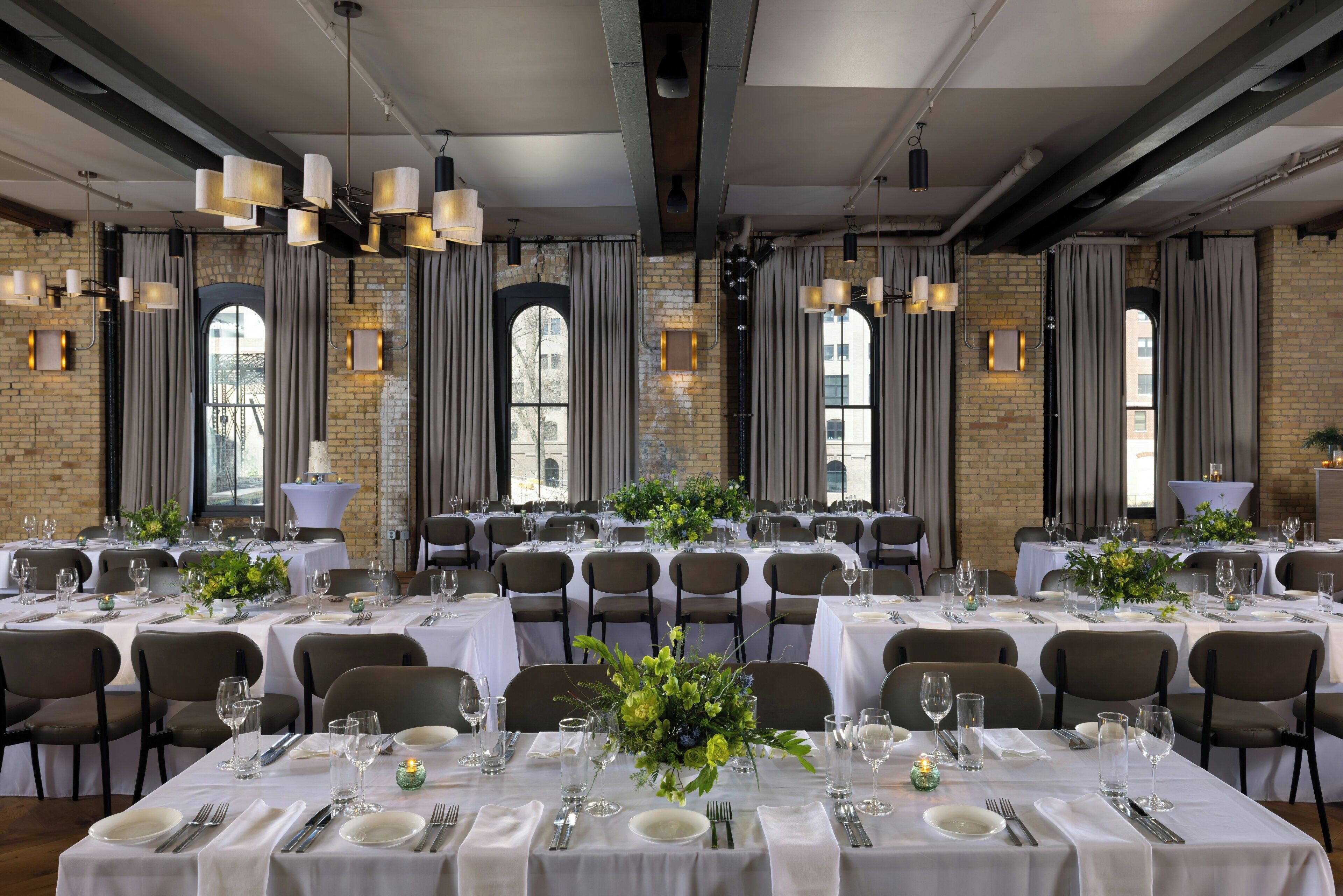 A banquet hall with multiple tables set for dining, featuring a brick wall and large windows.