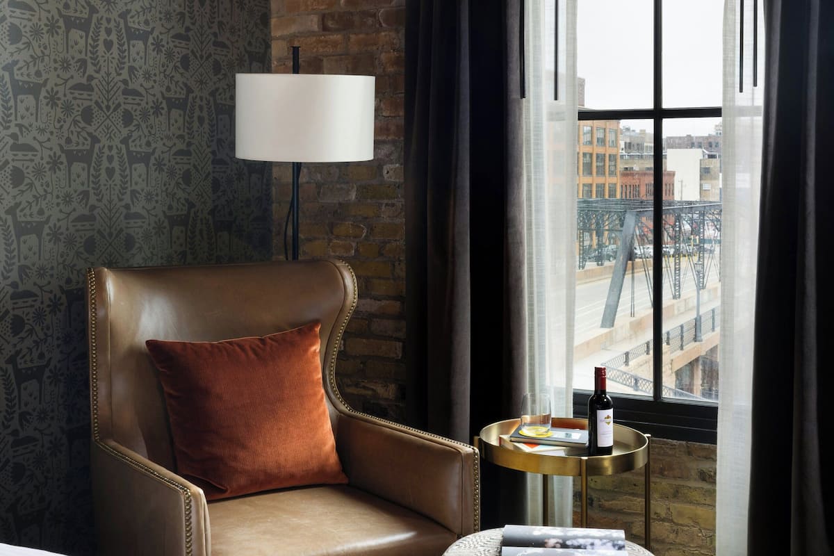 A hotel room with a bed, a leather armchair, a small round table with a lamp, a window with curtains, and a patterned wall.
