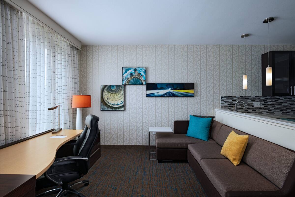 A hotel room with a desk, office chair, sofa, and a small kitchenette.