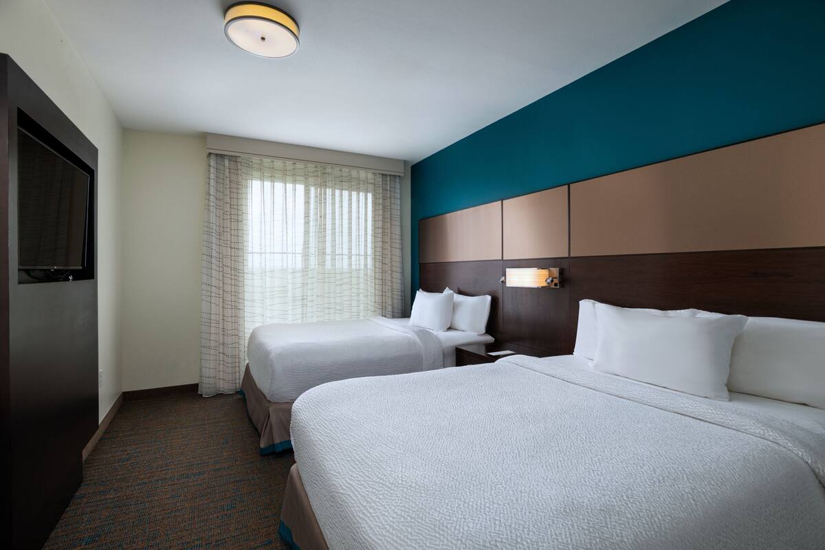 A hotel room with two beds, a TV, a window with curtains, and a headboard with a light fixture.