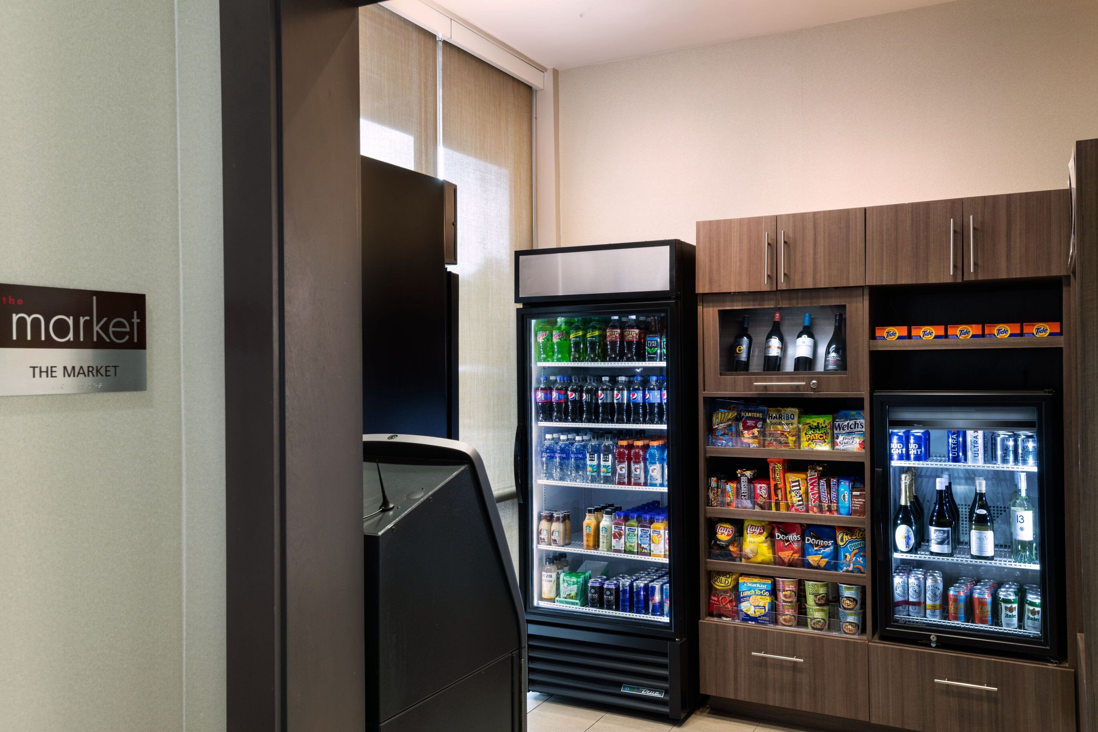 Habitación de hotel con minibar, un refrigerador lleno de bebidas, y una alacena con aperitivos y agua embotellada.