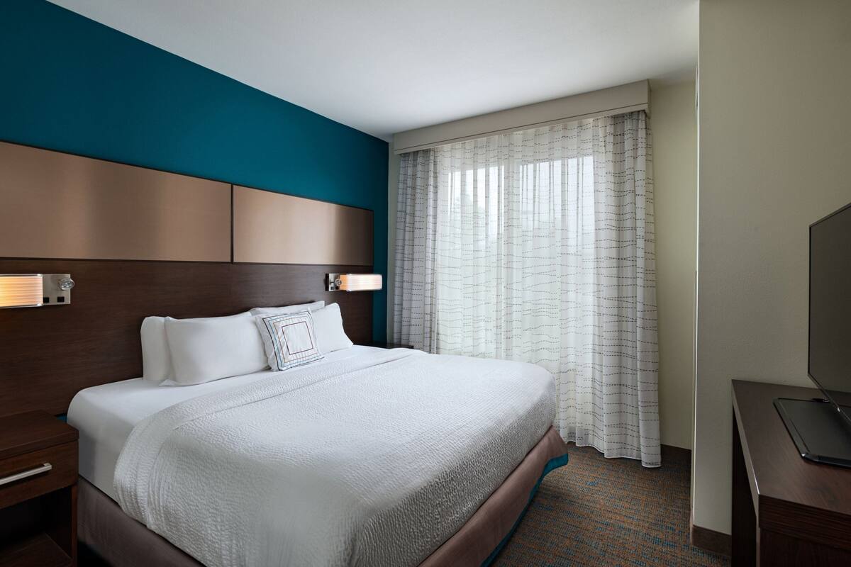 A hotel room with a large bed, a wooden headboard, a desk with a laptop, and a window with sheer curtains.
