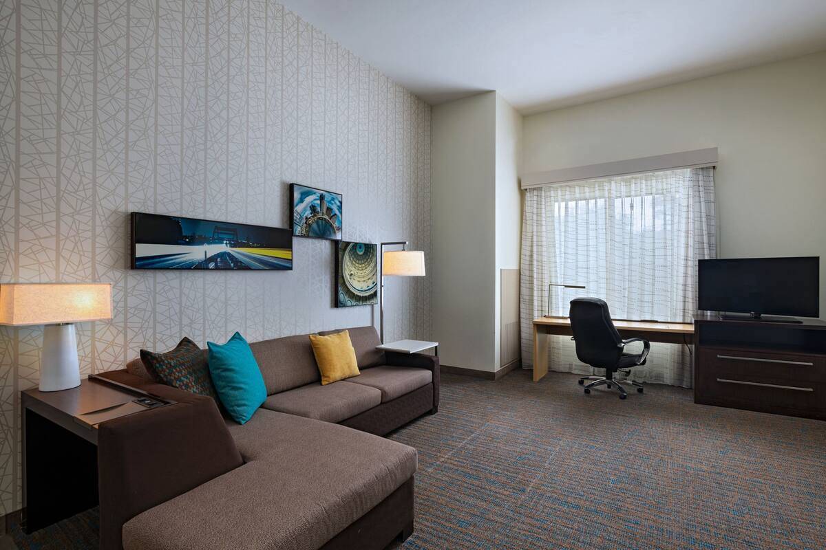 A hotel room with a sofa, a desk, a chair, a TV, and artwork on the wall.