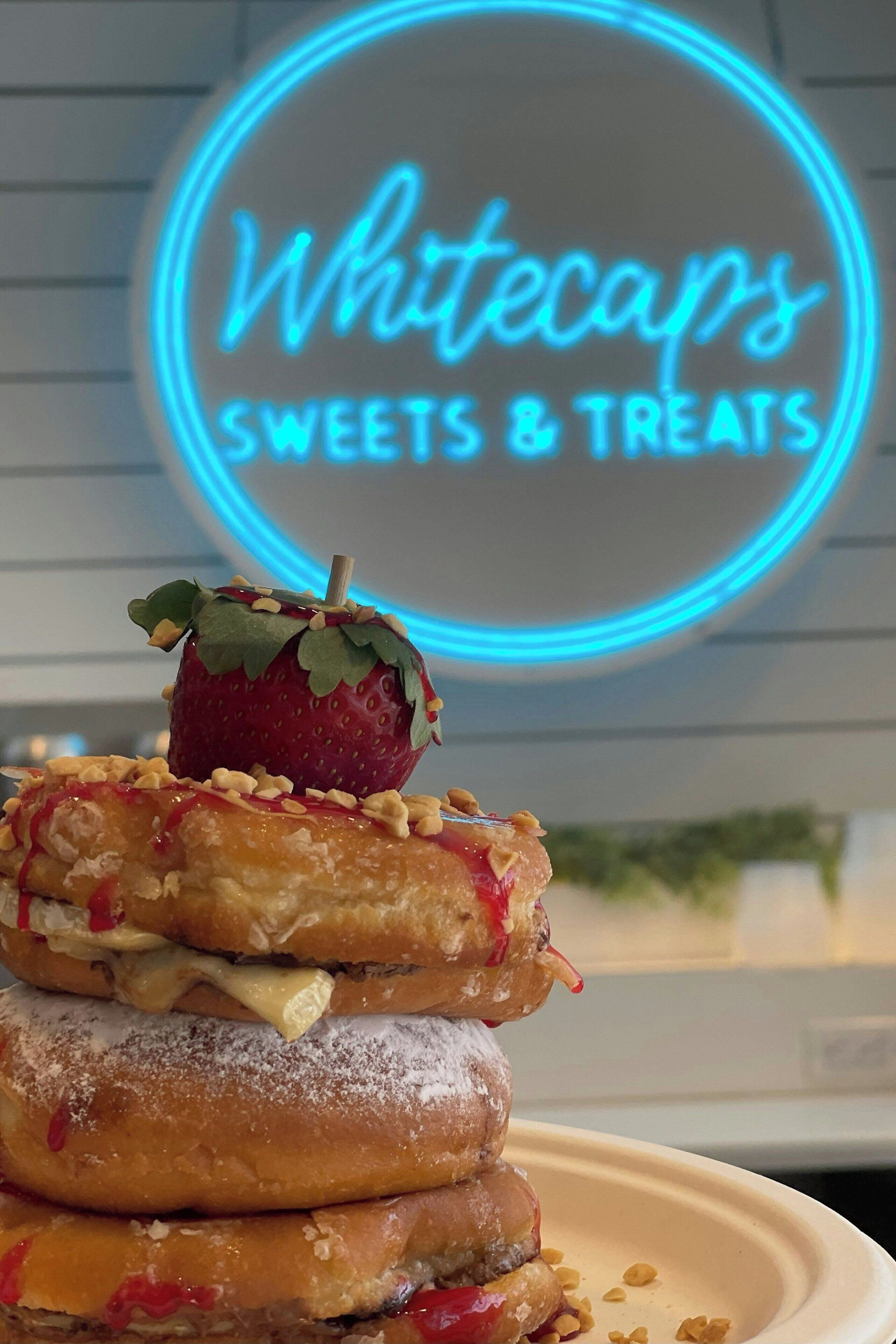 A stack of donuts with a neon sign in the background reading "Whitecaps Sweets & Treats".
