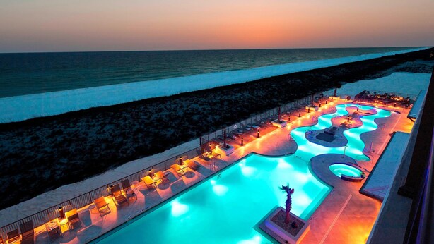 A swimming pool with a wavy design, surrounded by lounge chairs and a railing, adjacent to a beach.