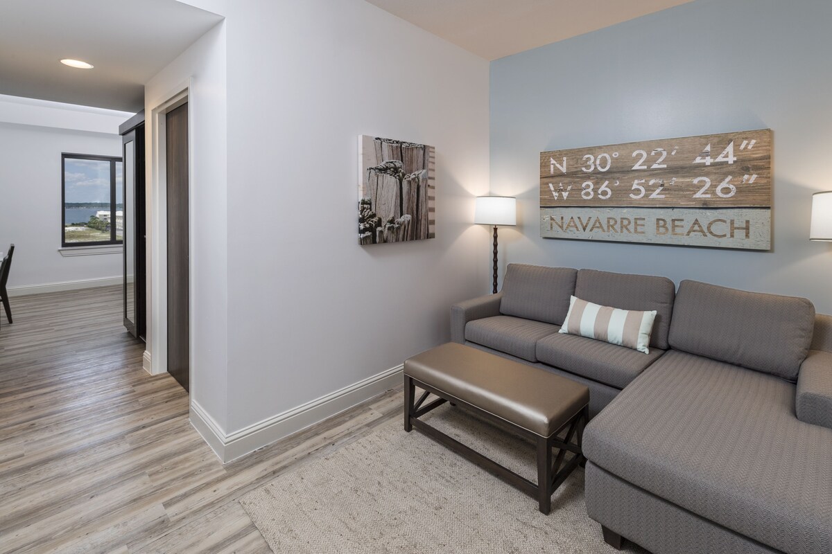 A modern living room with a grey sofa, a bench, and a wall-mounted lamp. A framed map of Navarre Beach is displayed on the wall.