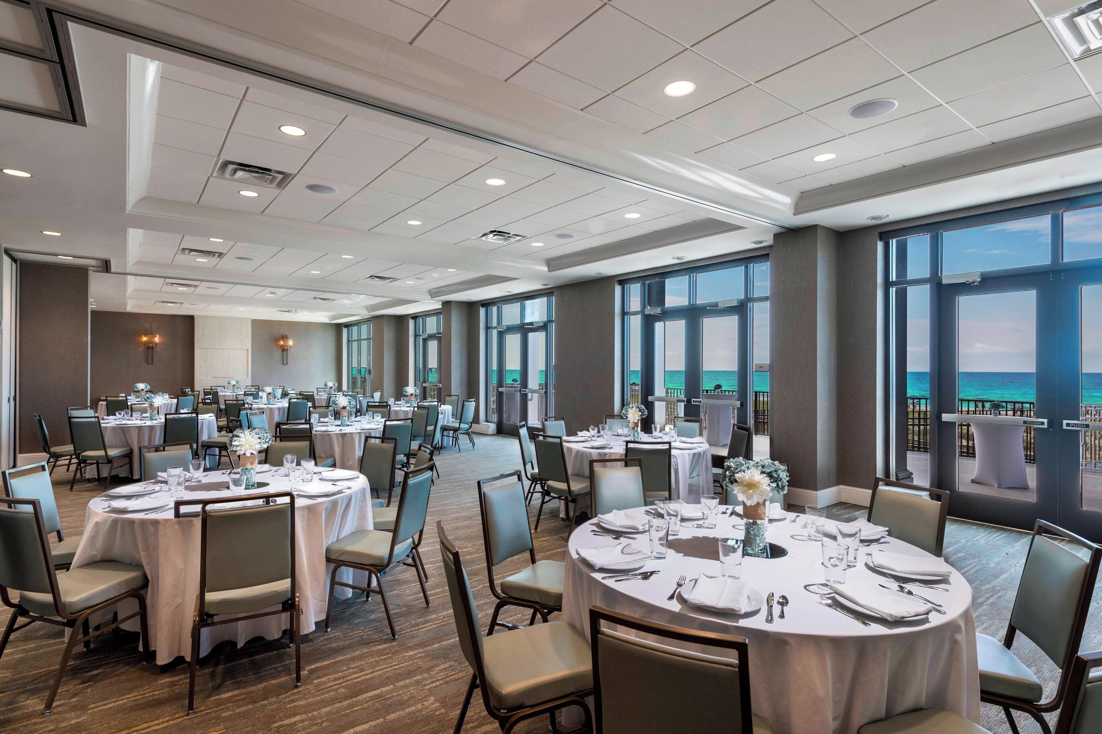A spacious banquet hall with round tables set for dining, large windows offering a view of the sea, and a modern ceiling design.