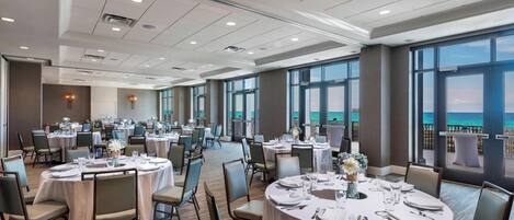 A spacious banquet hall with round tables set for dining, large windows offering a view of the sea, and a modern ceiling design.