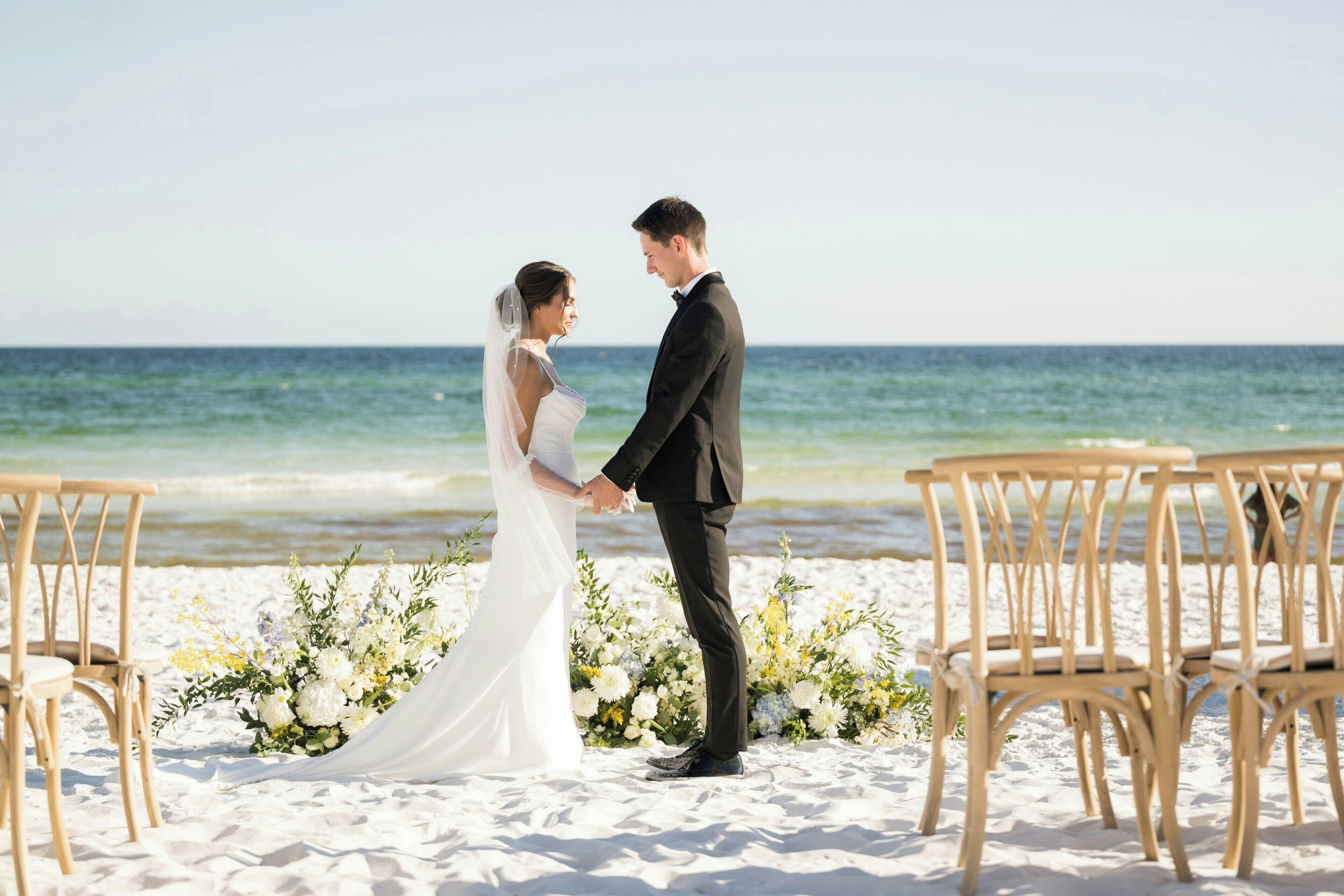 Ceremonia de boda en la playa, con una pareja tomándose de las manos, rodeada de arreglos florales y sillas de madera.