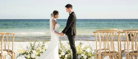 Ceremonia de boda en la playa, con una pareja tomándose de las manos, rodeada de arreglos florales y sillas de madera.