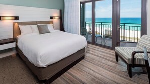 A hotel room with a bed, a chair, a view of the beach, and a balcony with a railing.