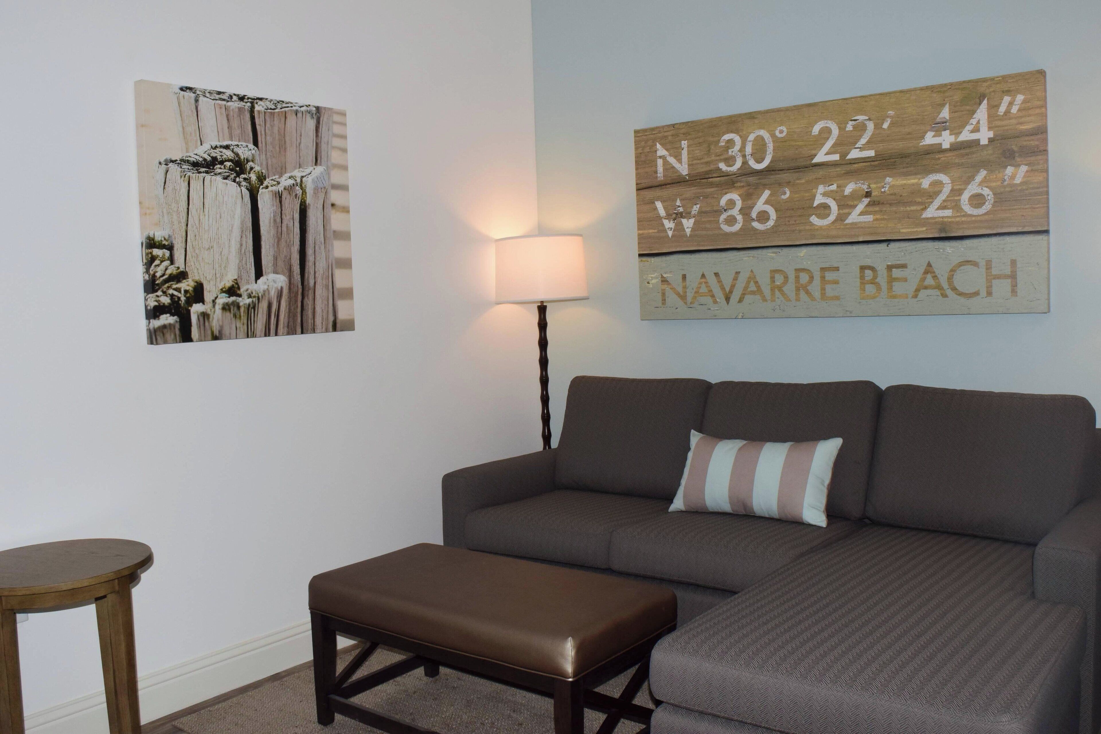A living room with a brown sofa, a brown leather ottoman, a wooden wall art piece, a lamp, and a wooden sign with coordinates and the text "NAVARRE BEACH".
