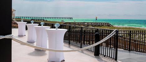 A balcony with white tables and chairs, a rope barrier, and a view of the ocean and pier.