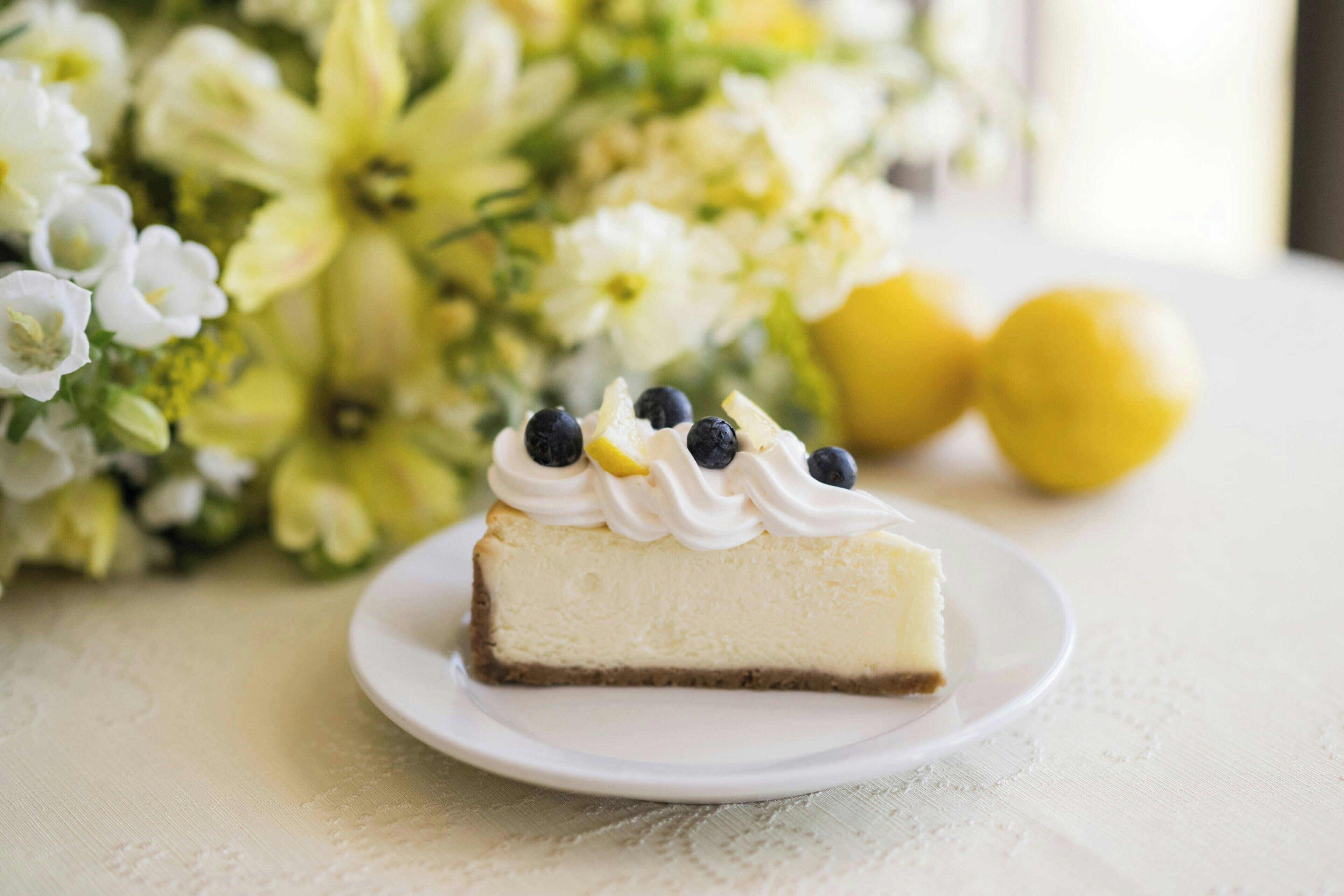 A slice of cheesecake on a white plate with blueberries and lemon on top, placed on a table with a floral arrangement in the background.