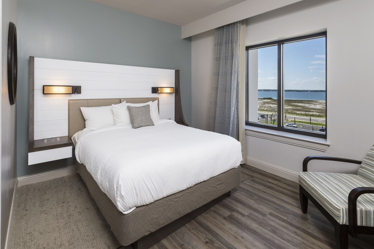 A modern hotel room with a large bed, a view of the beach, and a seating area.