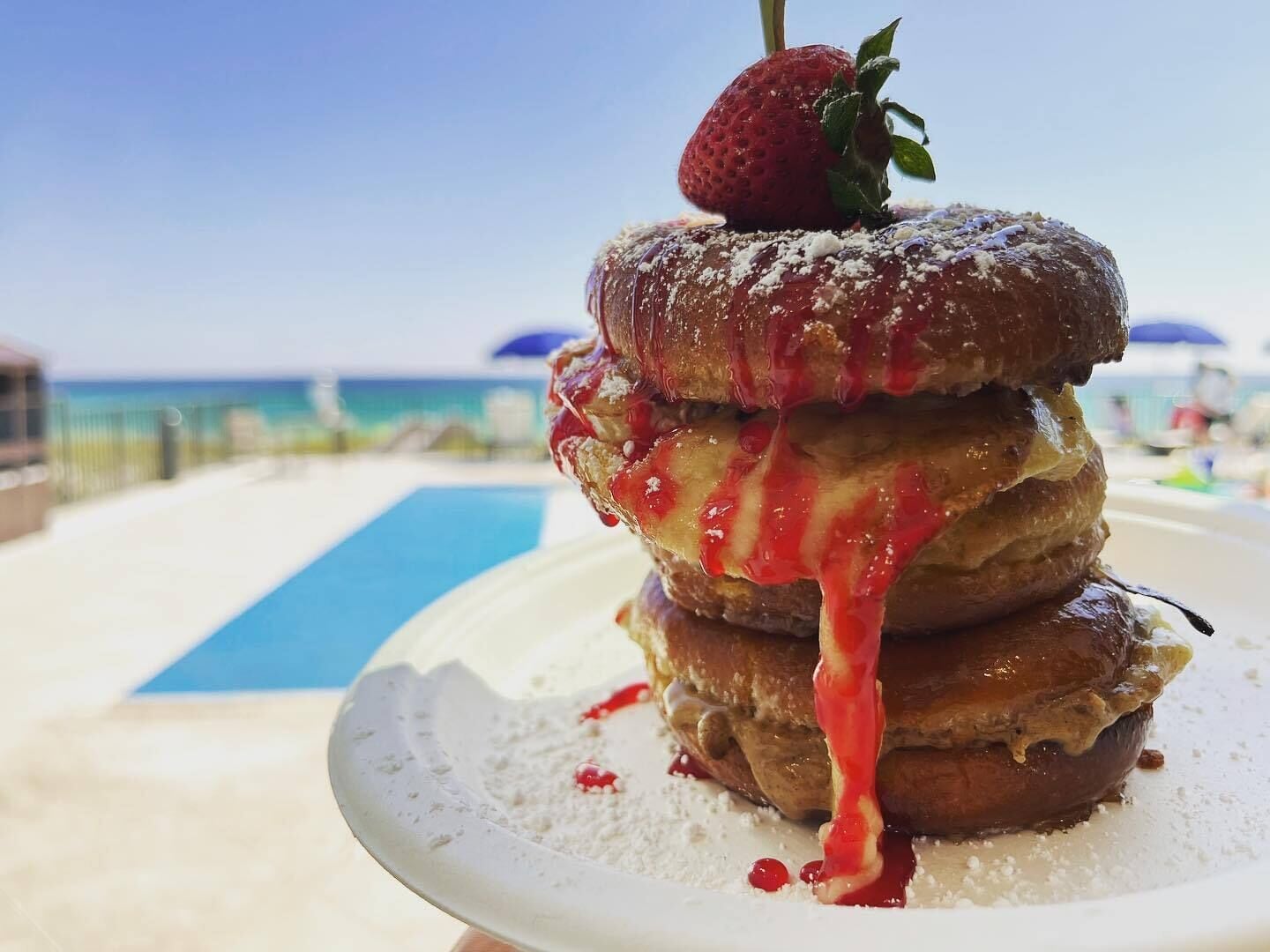 Una pila di pancake con una fragola sopra, servita su un piatto bianco con zucchero a velo e sciroppo rosso, su una spiaggia con una piscina sullo sfondo.