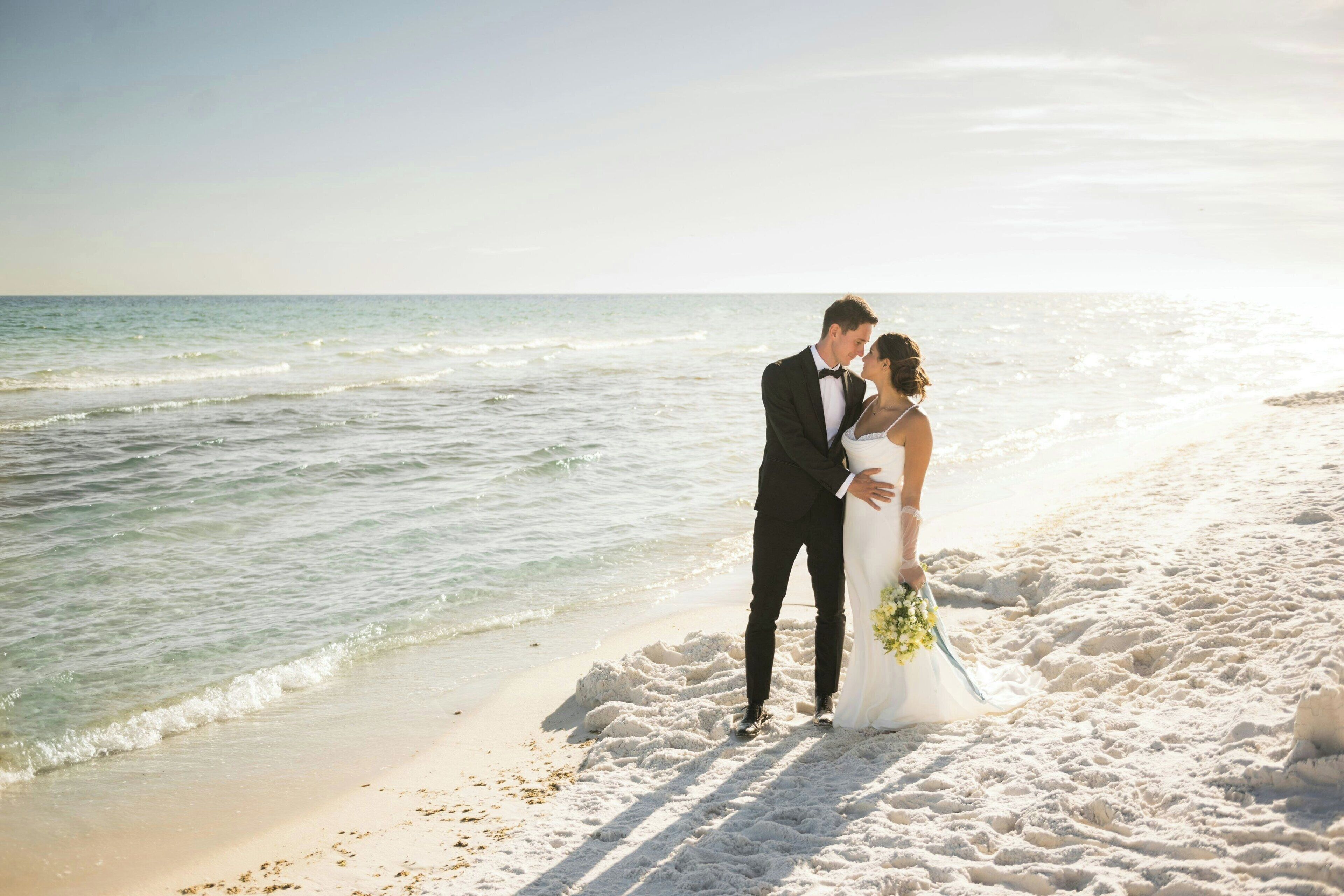 A beach wedding scene with a couple standing on the sand, facing the ocean.