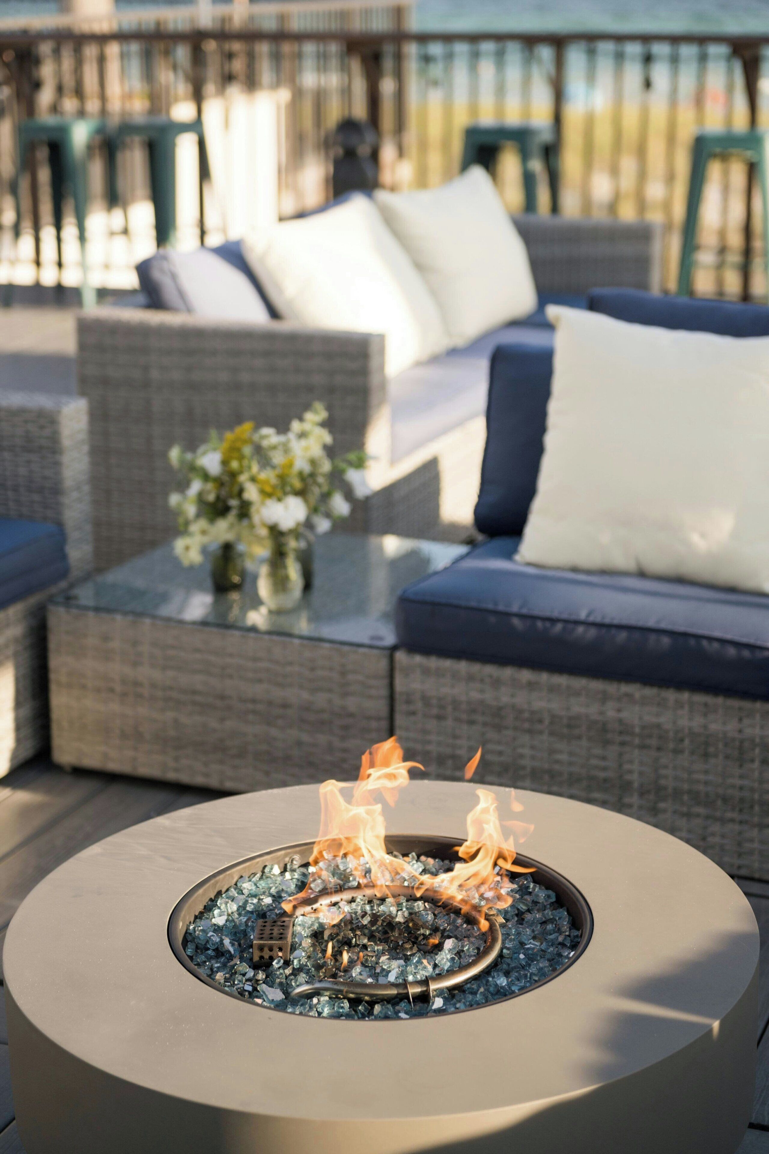 A modern outdoor seating area with a round fire pit in the center, surrounded by cushioned sofas and a glass coffee table with a vase of flowers.