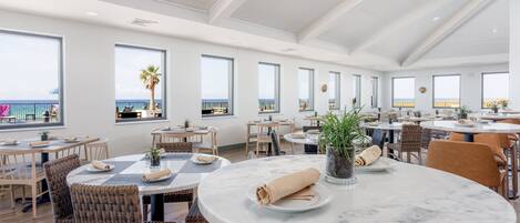 A spacious dining area with multiple round tables, wicker chairs, and large windows offering views of the beach.