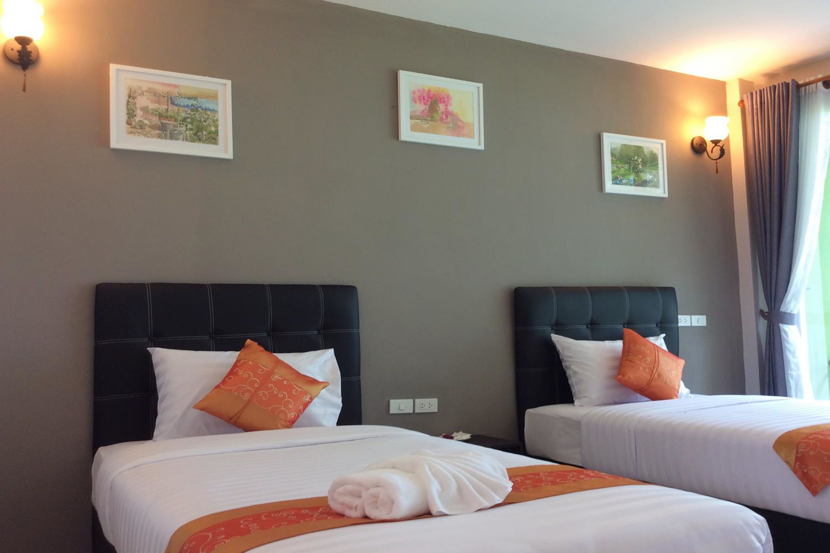 Habitación de hotel con dos camas, cada una con cabecero negro, ropa de cama blanca y almohadas decorativas naranjas. Hay cuadros enmarcados en la pared y una ventana con cortinas.