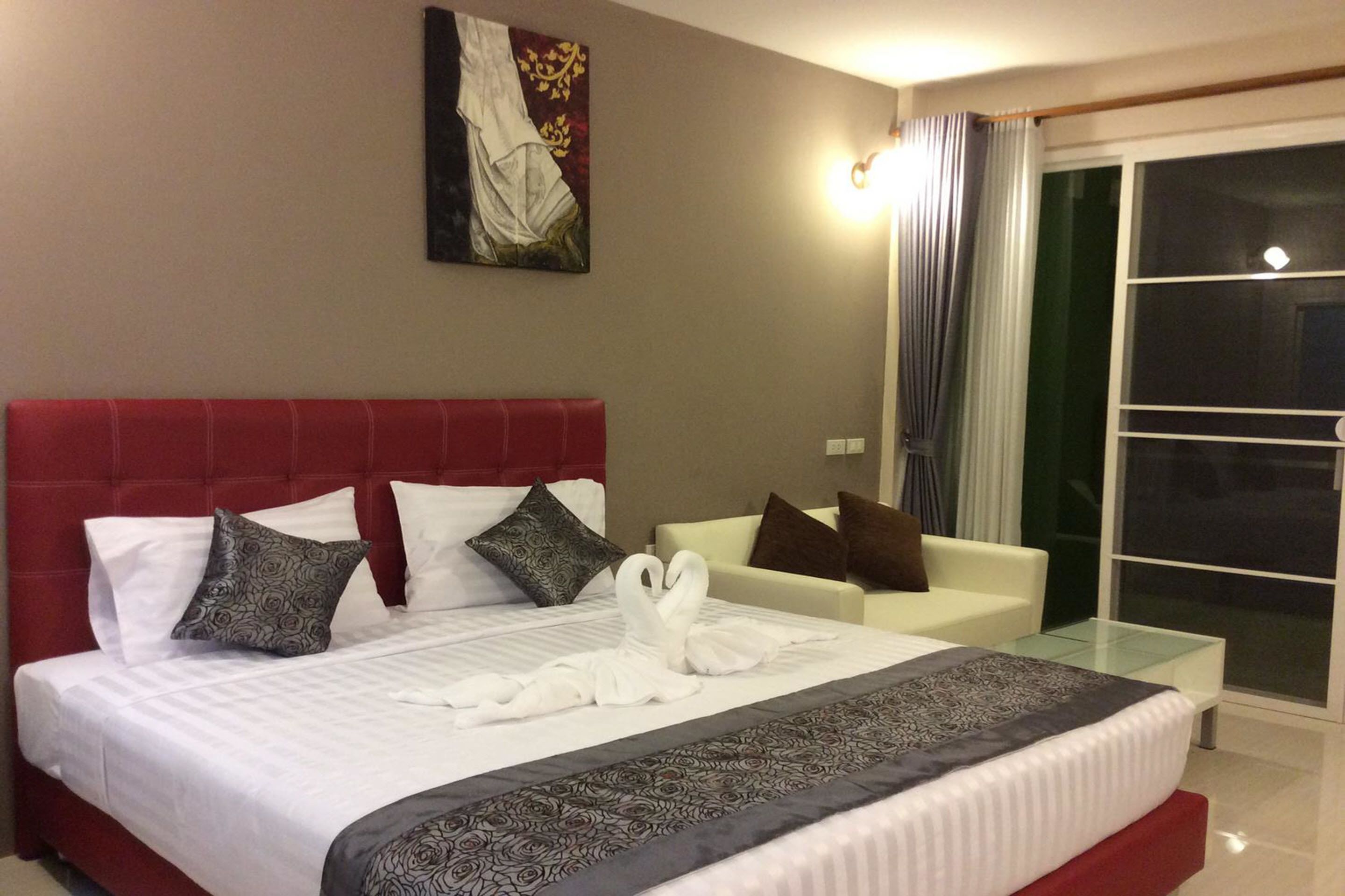 A hotel room with a large bed, a red headboard, a glass coffee table, a white sofa, and a painting on the wall.